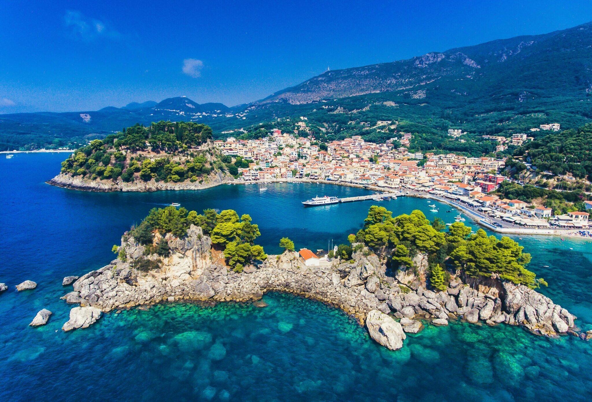 Vue aérienne d’une baie méditerranéenne avec un îlot rocheux, une ville côtière aux toits orangés et des eaux turquoise cristallines