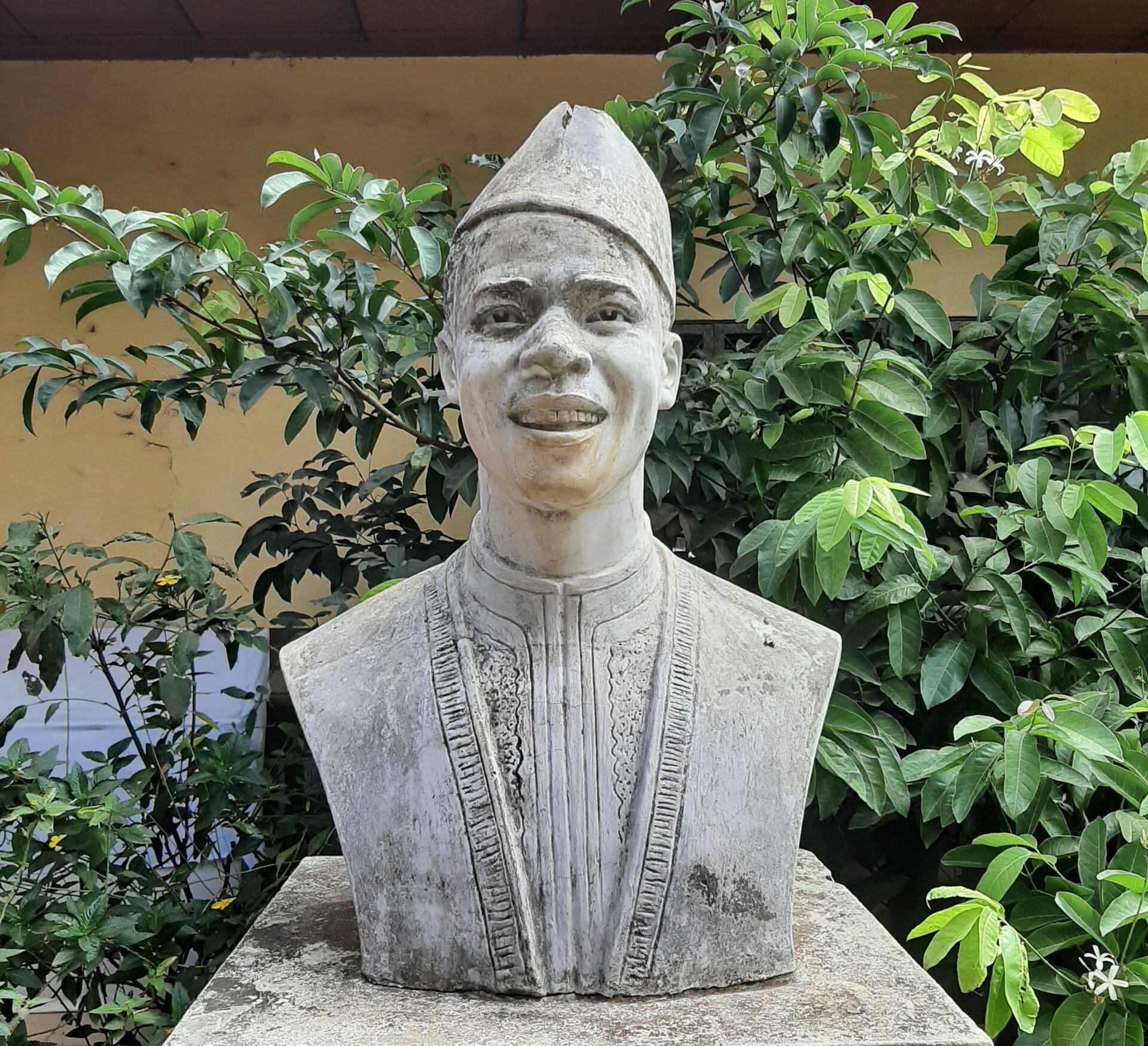 Buste en pierre sculptée vêtu de tunique traditionnelle, entouré de verdure luxuriante dans un jardin