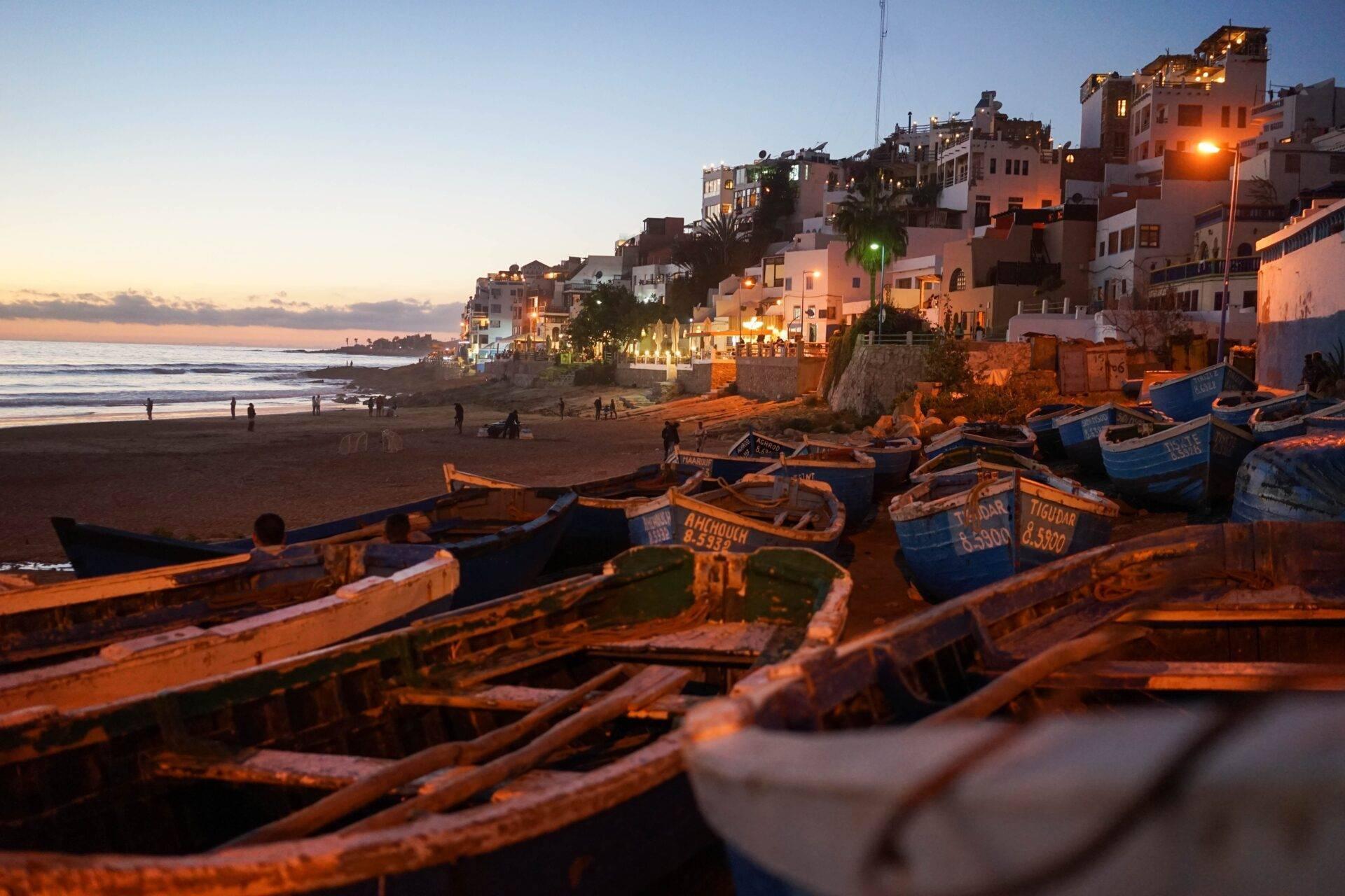 Des barques de pêche colorées sur une plage au coucher du soleil, face à des maisons blanches perchées sur la falaise