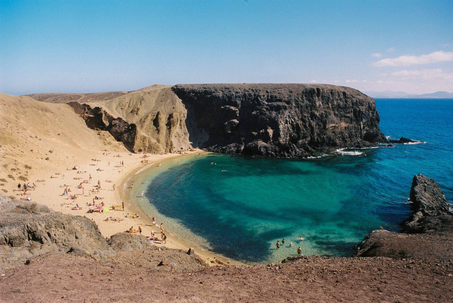 Une plage de sable doré encadrée de falaises noires et une mer turquoise cristalline avec des baigneurs