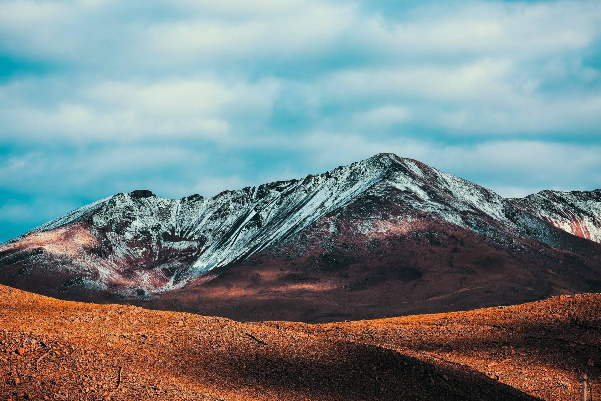 Un volcan aux pentes multicolores recouvertes de neige dominan un désert rocheux aride sous un ciel dégagé