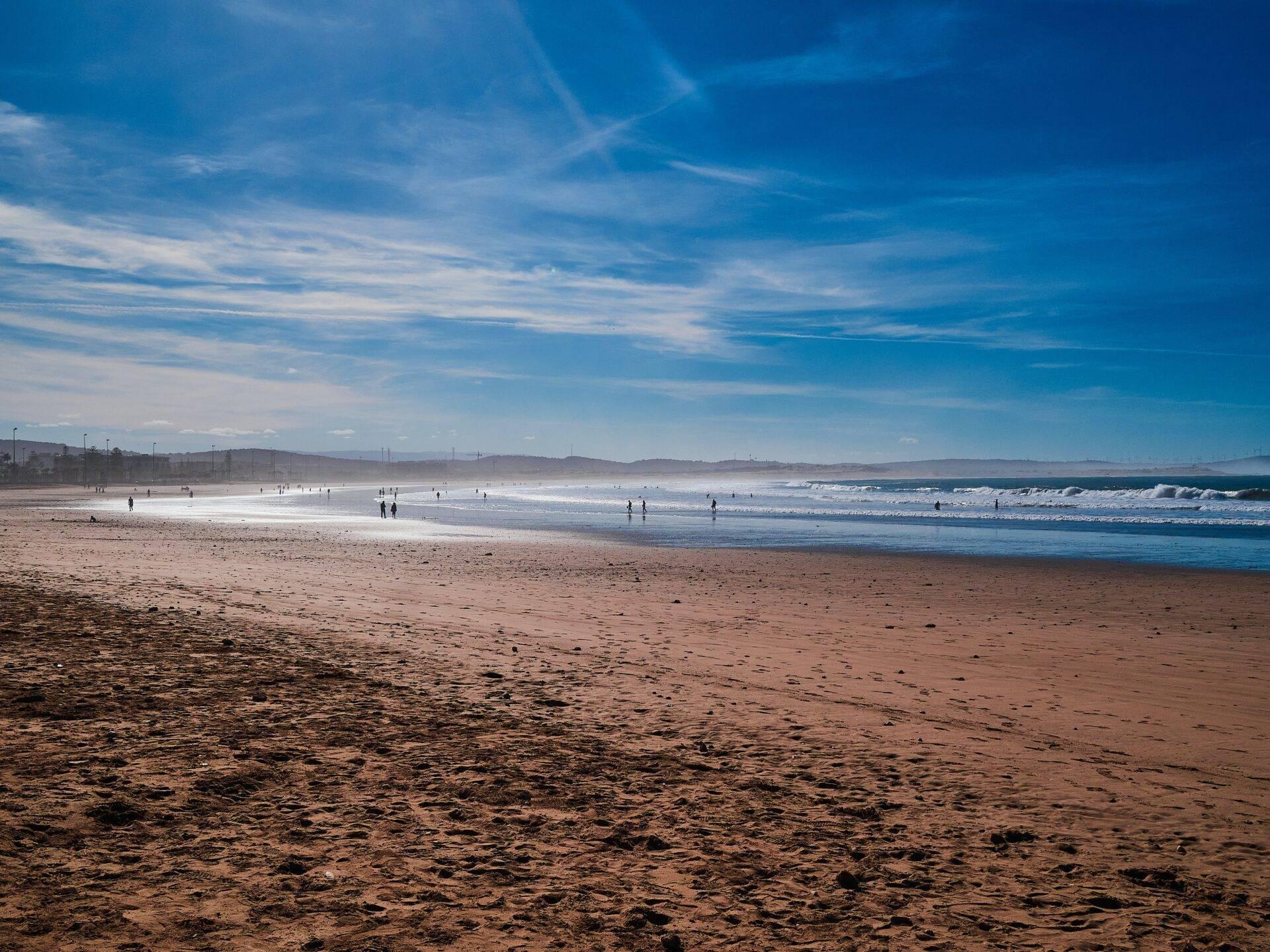 Une plage de sable fin avec des baigneurs, des vagues et des montagnes en arrière-plan sous un ciel bleu dégagé