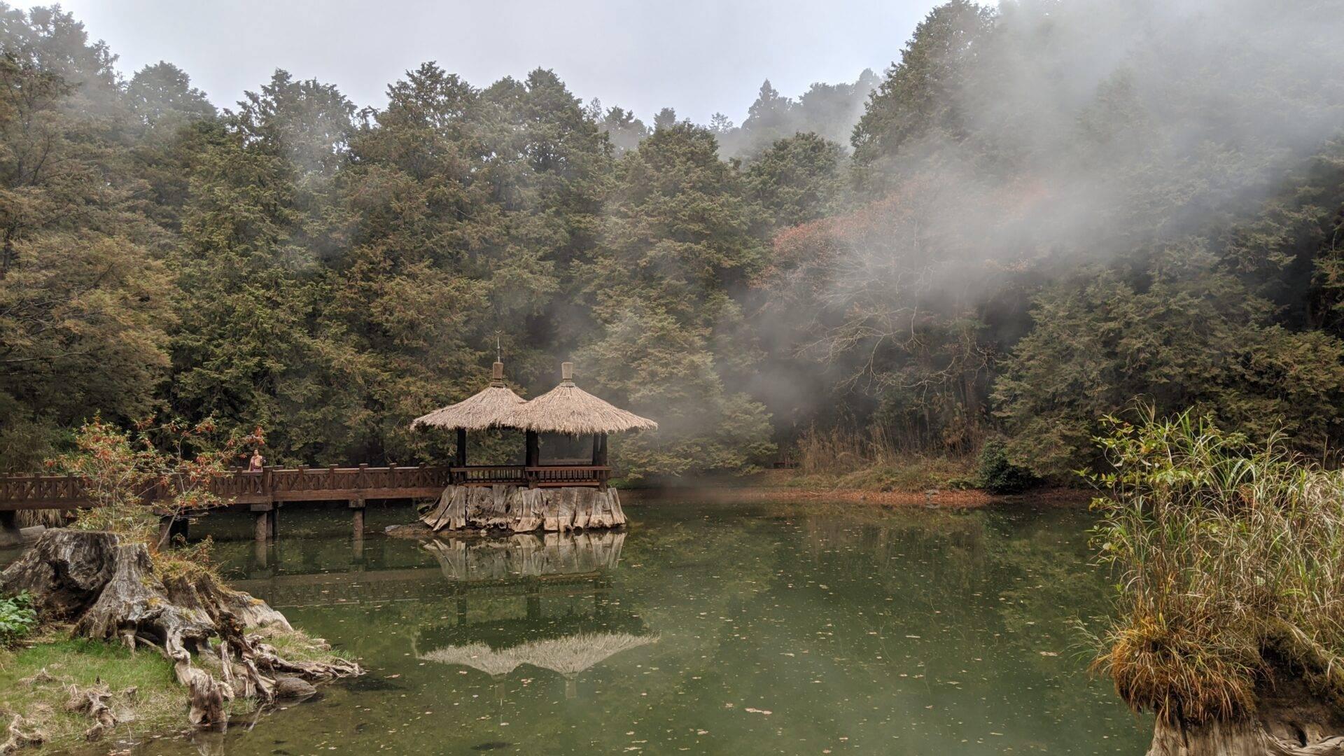 Pavillon traditionnel avec toits de chaume sur un lac entouré de forêts montagneuses dans la brume matinale