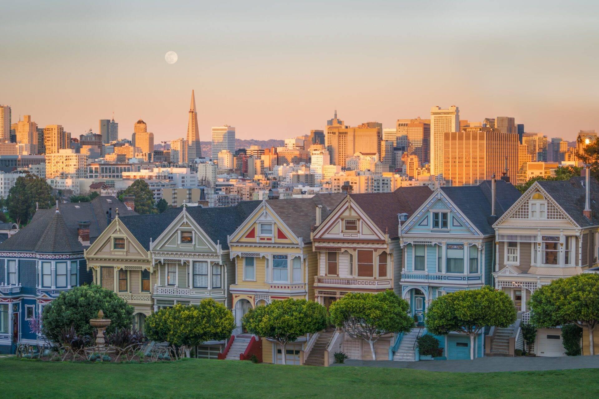 Rangée de maisons victoriennes colorées face à un parc avec skyline urbaine en arrière-plan au coucher du soleil