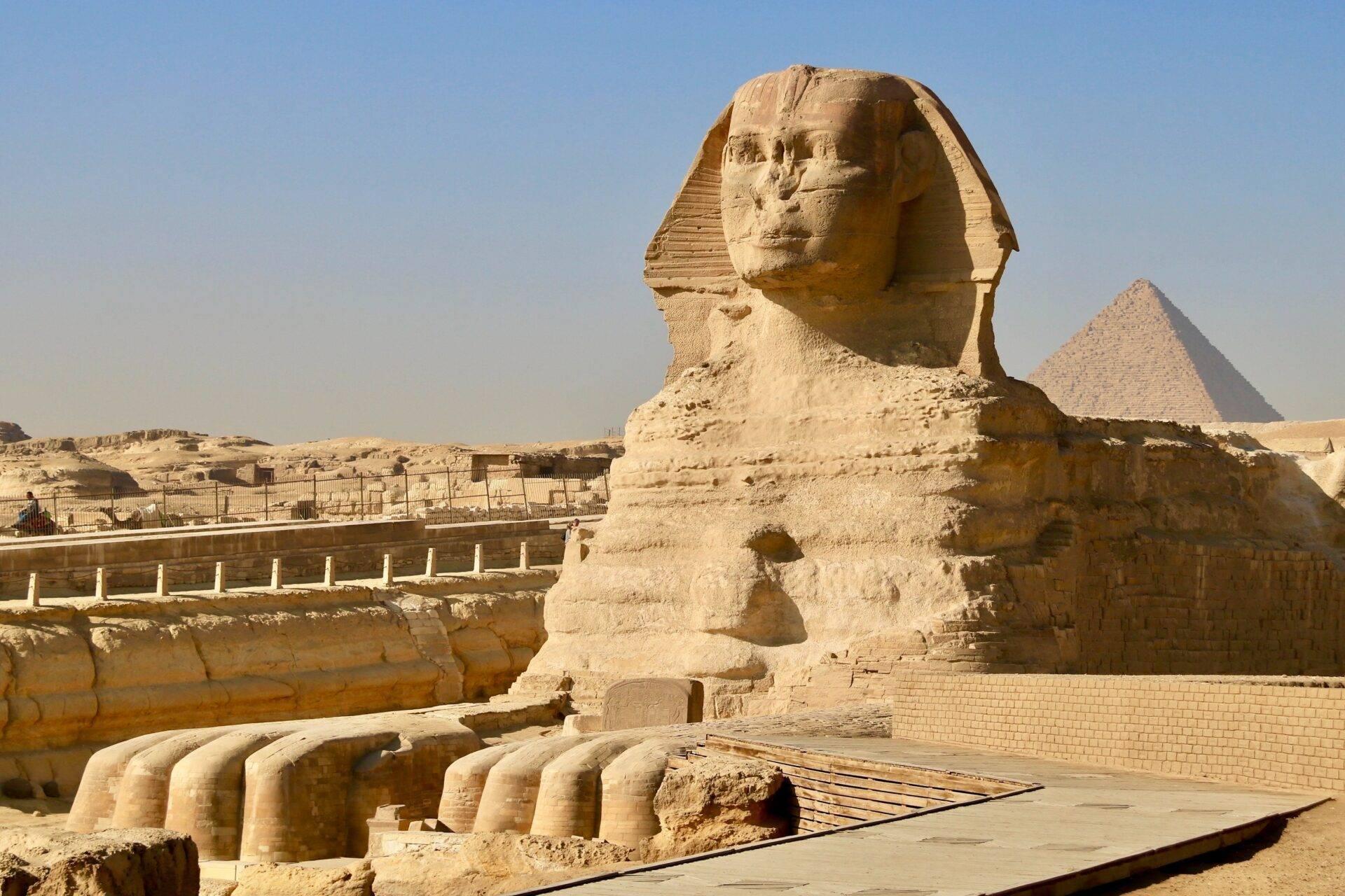 Le Grand Sphinx de Gizeh face à une pyramide, avec des vestiges architecturaux et des barrières de protection