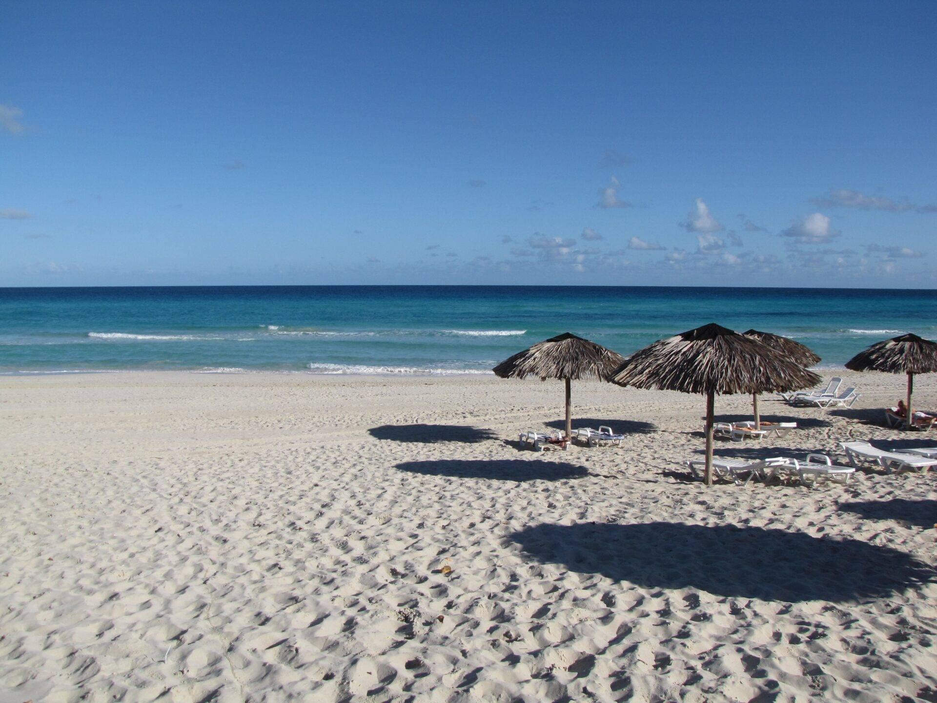 Une plage de sable blanc avec parasols en paille, chaises longues et mer turquoise sous un ciel bleu dégagé