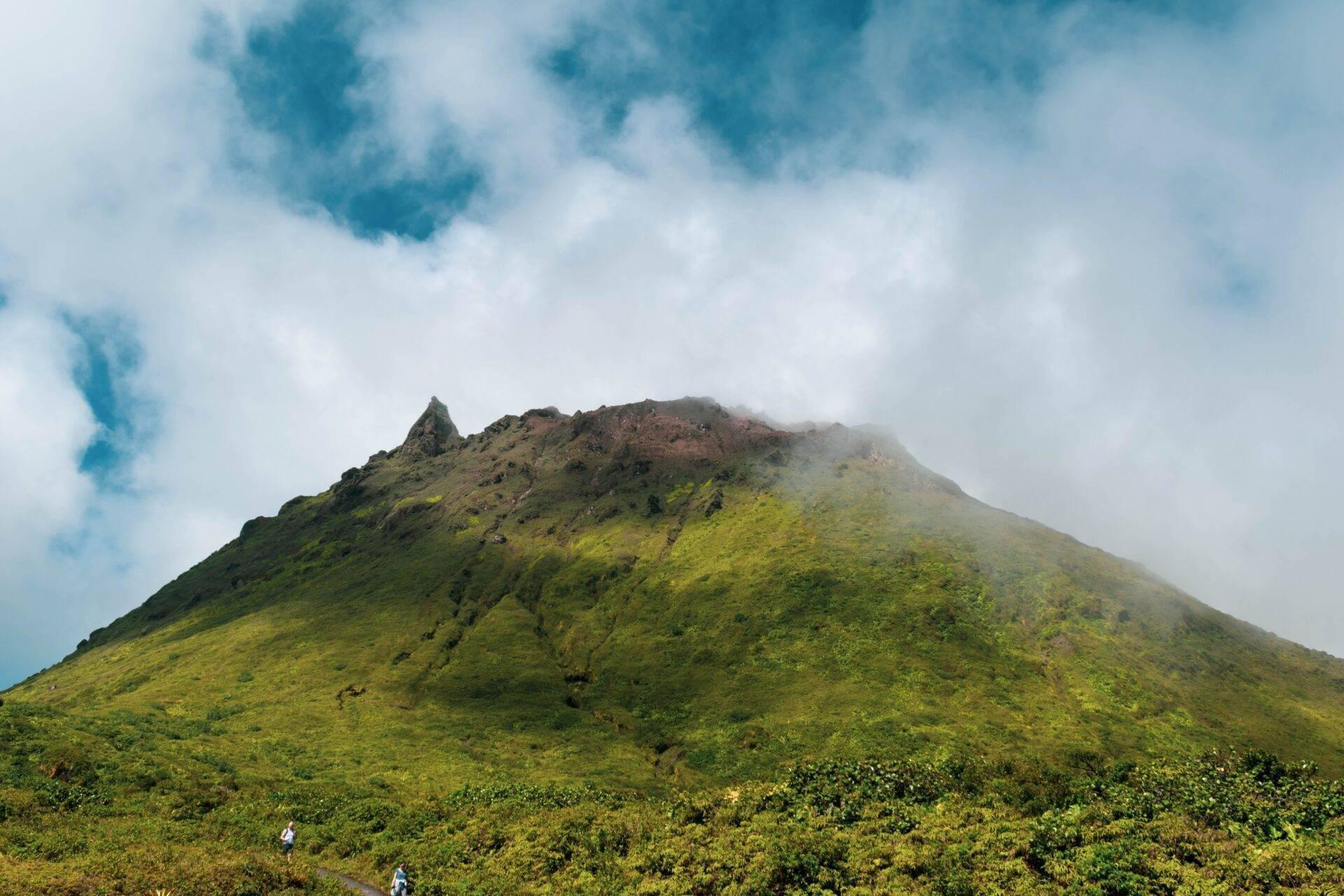 Un volcan conique couvert de végétation luxuriante avec son sommet partiellement enveloppé de nuages sous un ciel bleu