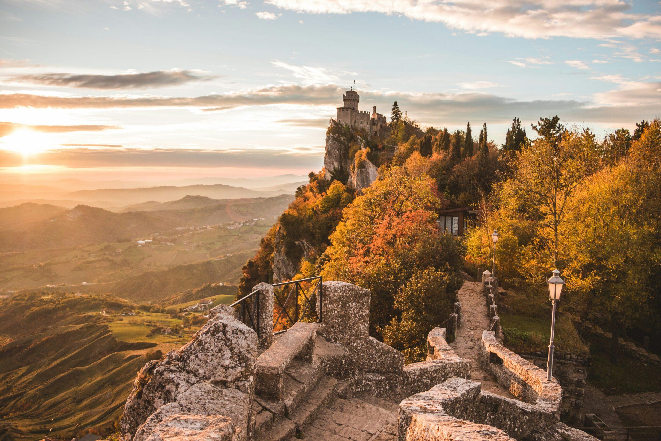 Vue depuis une terrasse de pierre sur un château médiéval perché sur une falaise rocheuse, entouré de forêts automnales dorées