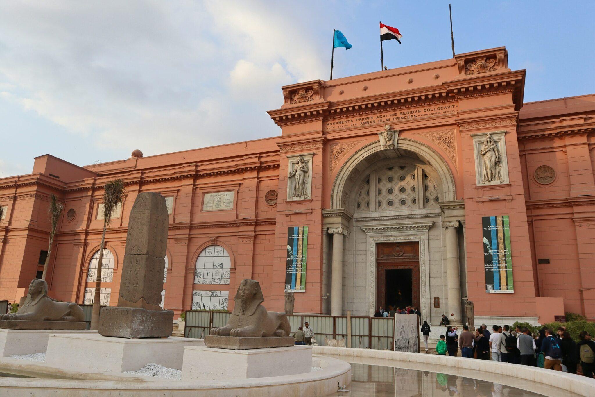 Façade rose d’un musée égyptien avec statues de sphinx, obelisque et drapeaux, visiteurs à l’entrée