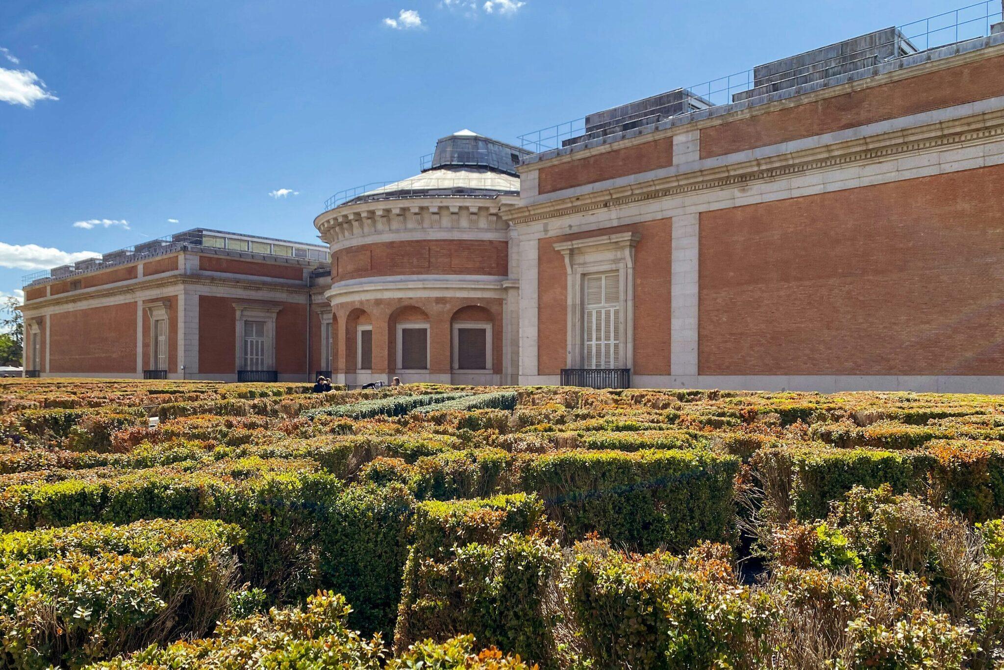 Musée avec façade en brique rouge et dôme, entouré d’un jardin de buis en topiaire géométrique sous ciel bleu