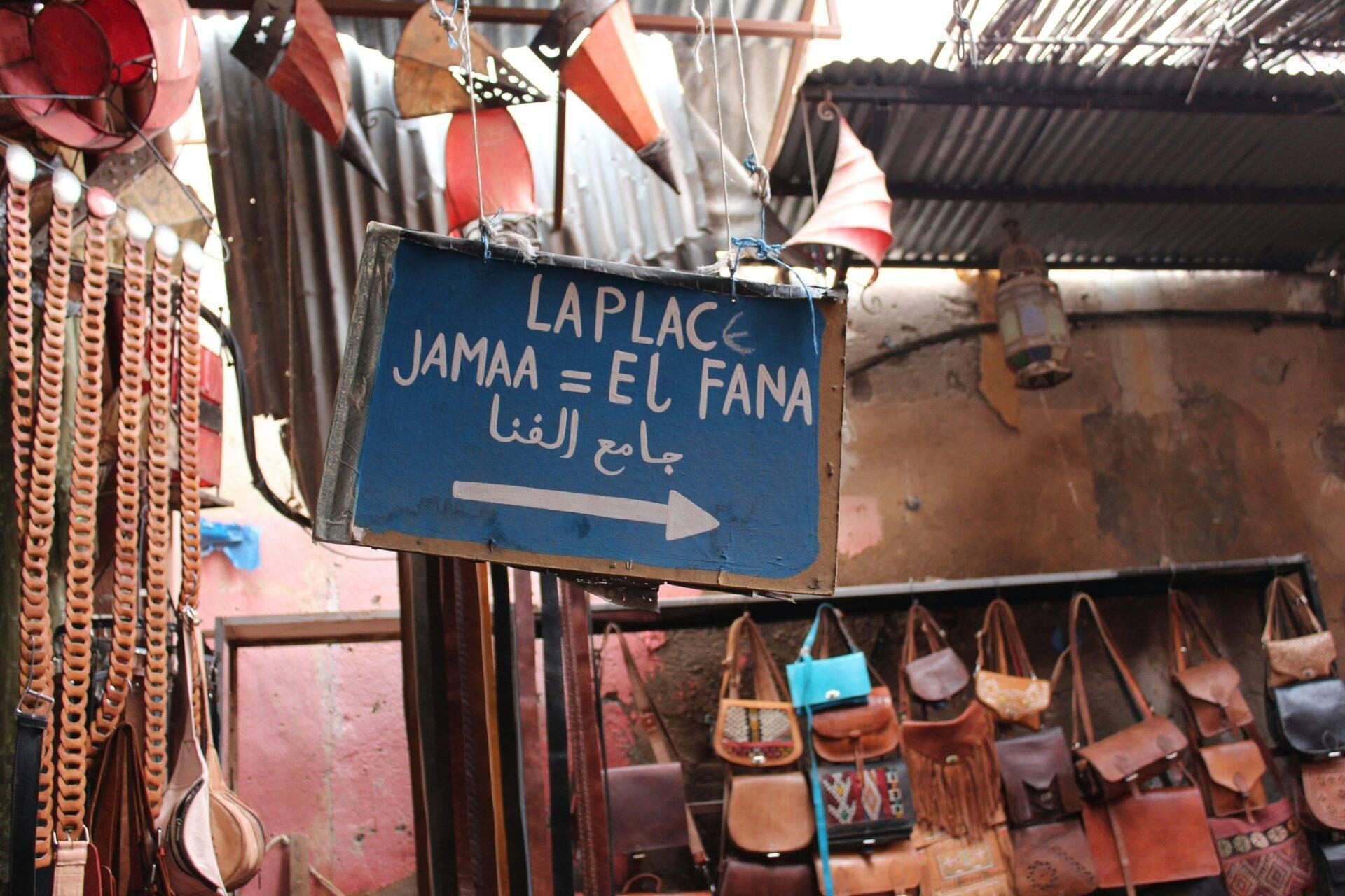 Panneau directional bleu en français et arabe pointant vers une boutique de maroquinerie avec sacs en cuir exposés dans une médina marocaine