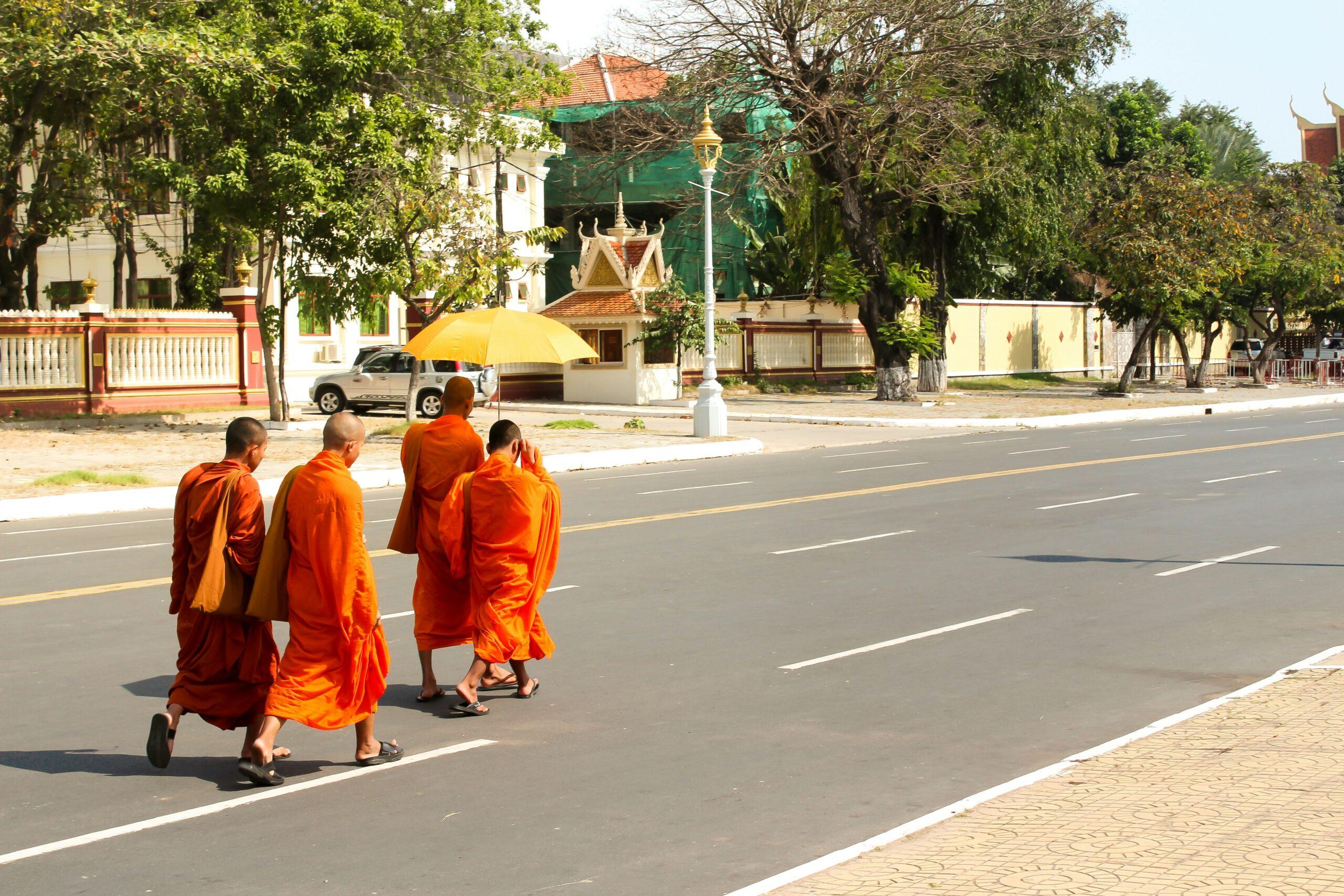 Quatre moines en robes orange traversent une large avenue ensoleillée devant un temple bouddhiste traditionnel