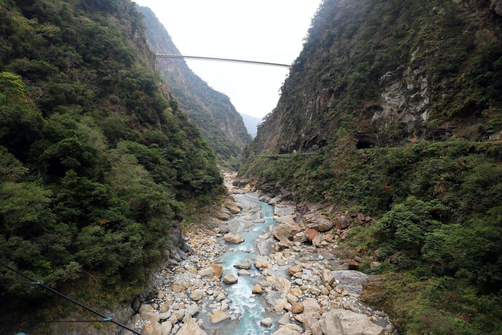 Une gorge montagneuse profonde traversée par une rivière turquoise, encadrée de falaises verdoyantes et d’un pont suspendu