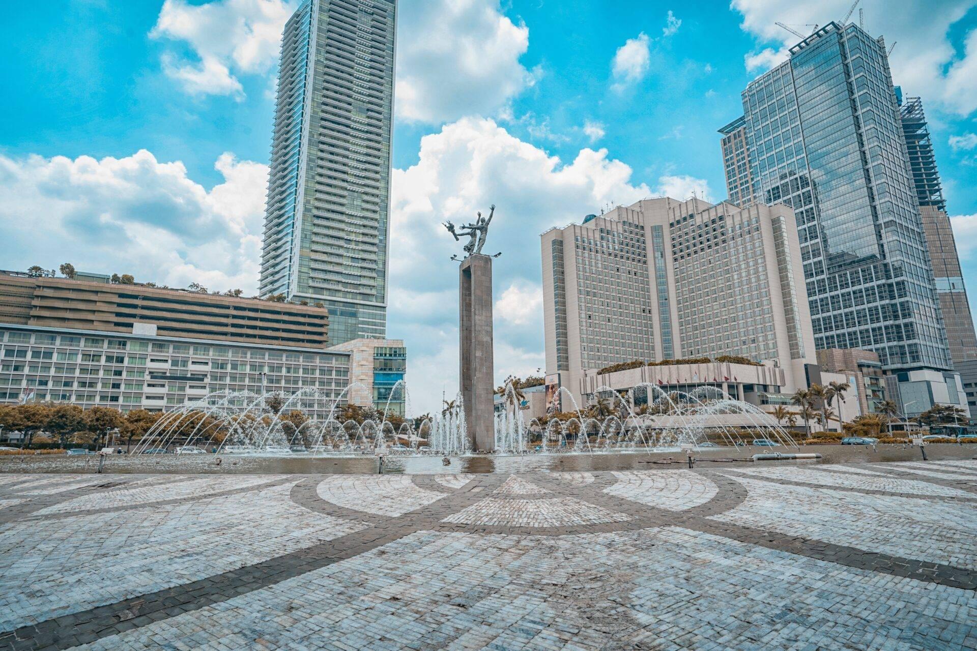 Une place urbaine pavée avec une statue monumentale, fontaines jaillissantes et gratte-ciels modernes sous ciel bleu