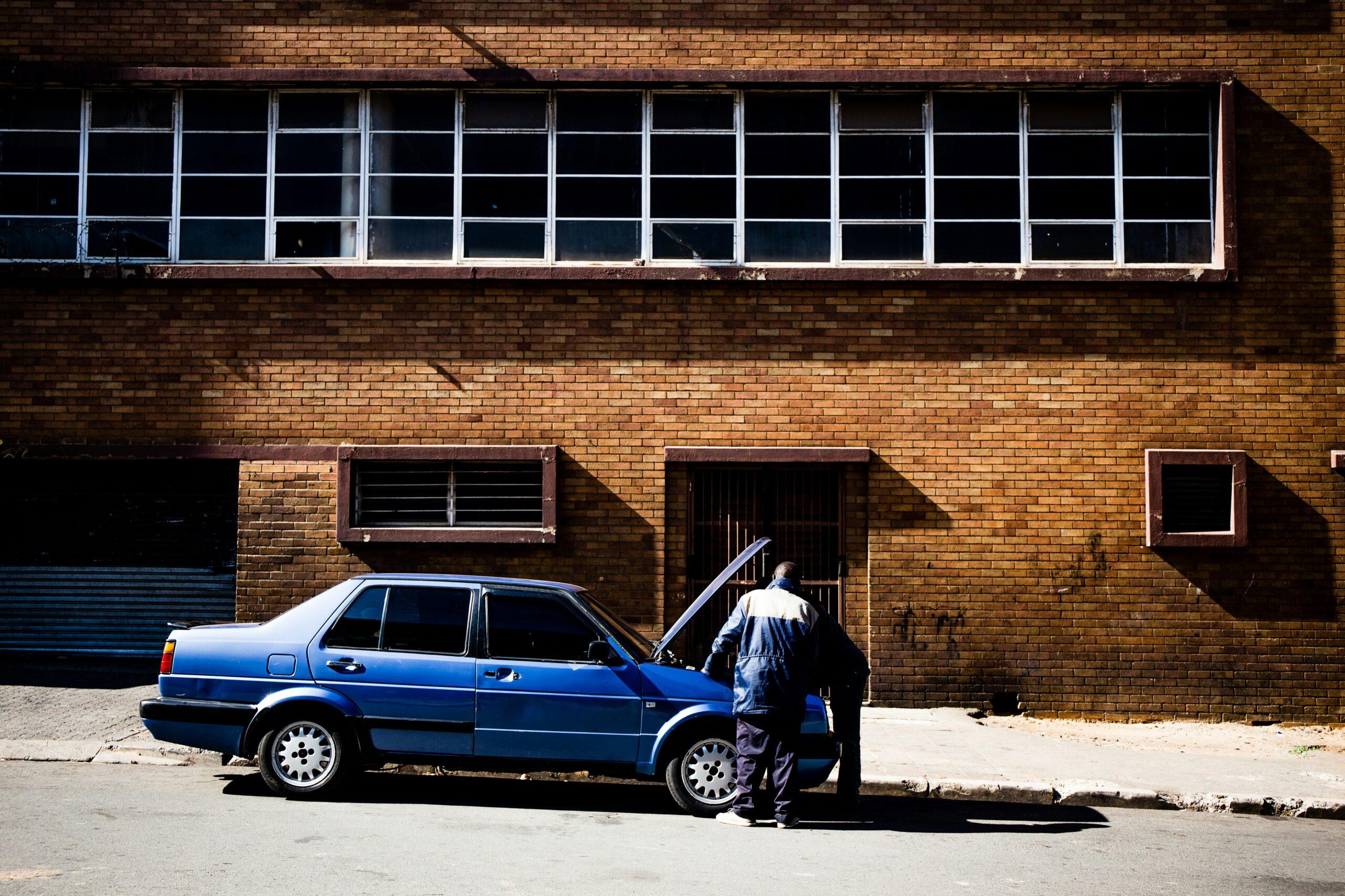 Un mécanicien répare une voiture bleue devant un bâtiment industriel en brique avec fenêtres grillagées