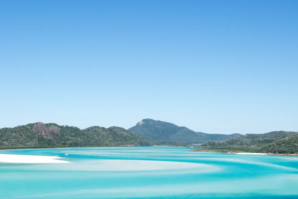 Lagon turquoise avec plage de sable blanc, îles boisées et montagne verdoyante sous ciel bleu dégagé