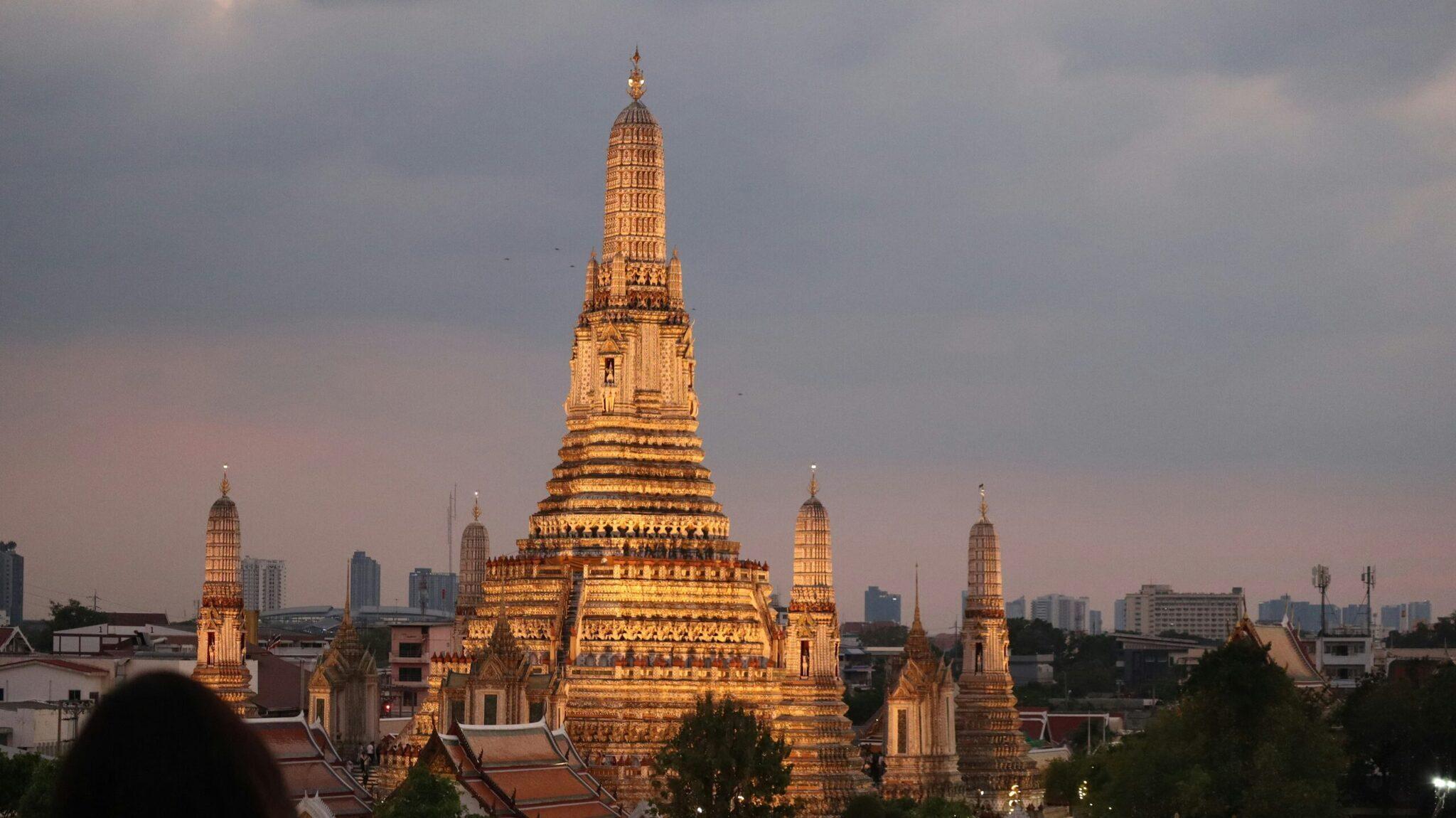 Un grand temple bouddhiste thaïlandais aux toits dorés illuminés au coucher du soleil, entouré de la skyline urbaine