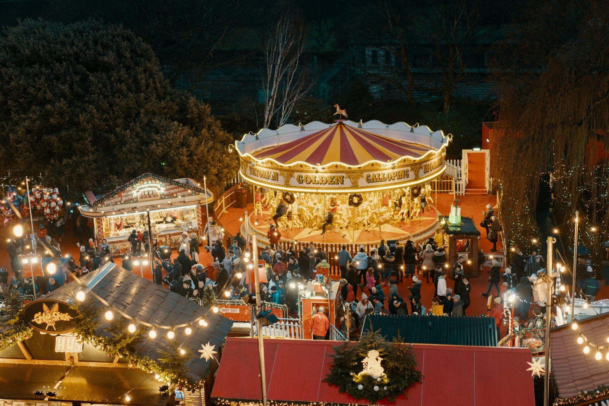 Un marché de Noël nocturne avec un carrousel illuminé doré et des stands décorés, foule visiteurs sous guirlandes lumineuses