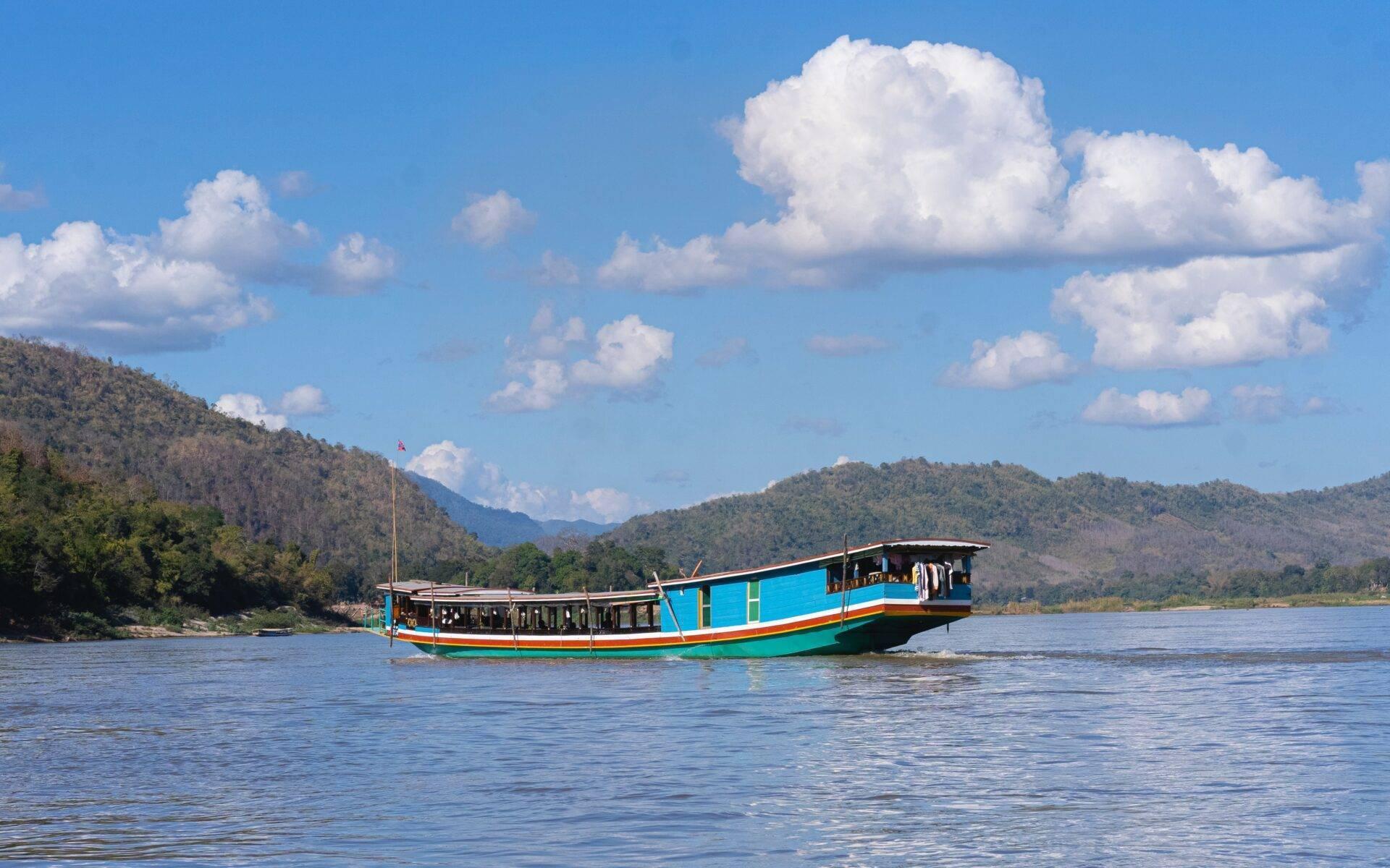 Un bateau touristique bleu traditionnel navigue sur un fleuve bordé de montagnes boisées sous un ciel nuageux
