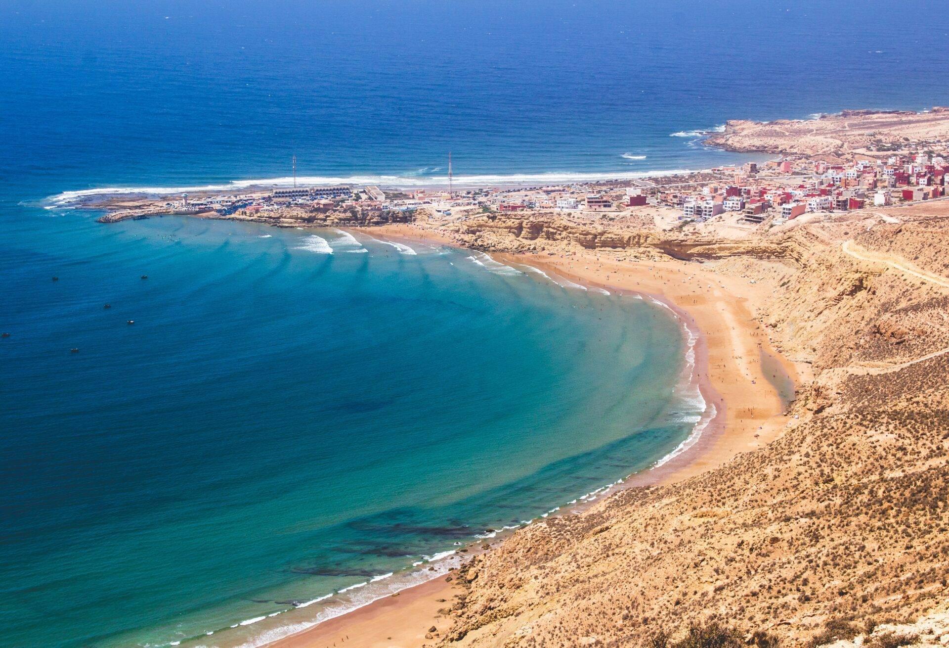 Vue aérienne d’une baie côtière marocaine avec plage de sable, ville bâtie en arrière-plan et eaux turquoise cristallines