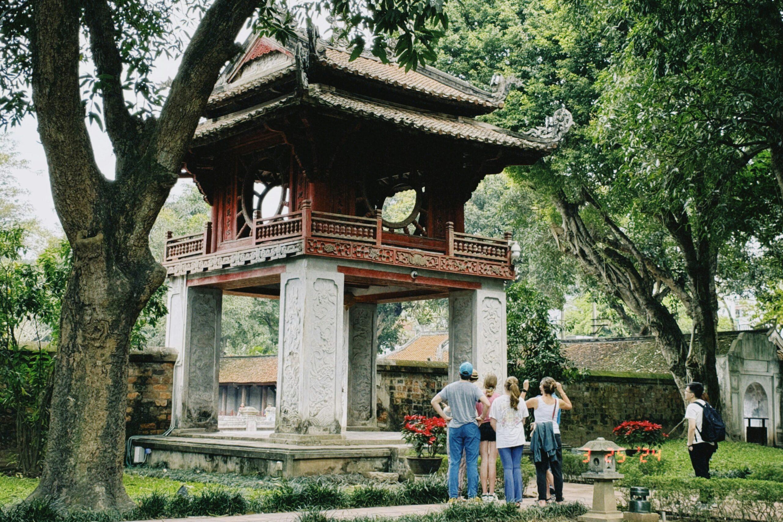 Un pavillon traditionnel asiatique aux toits recourbés entouré de grands arbres anciens, avec des visiteurs admirant l’architecture