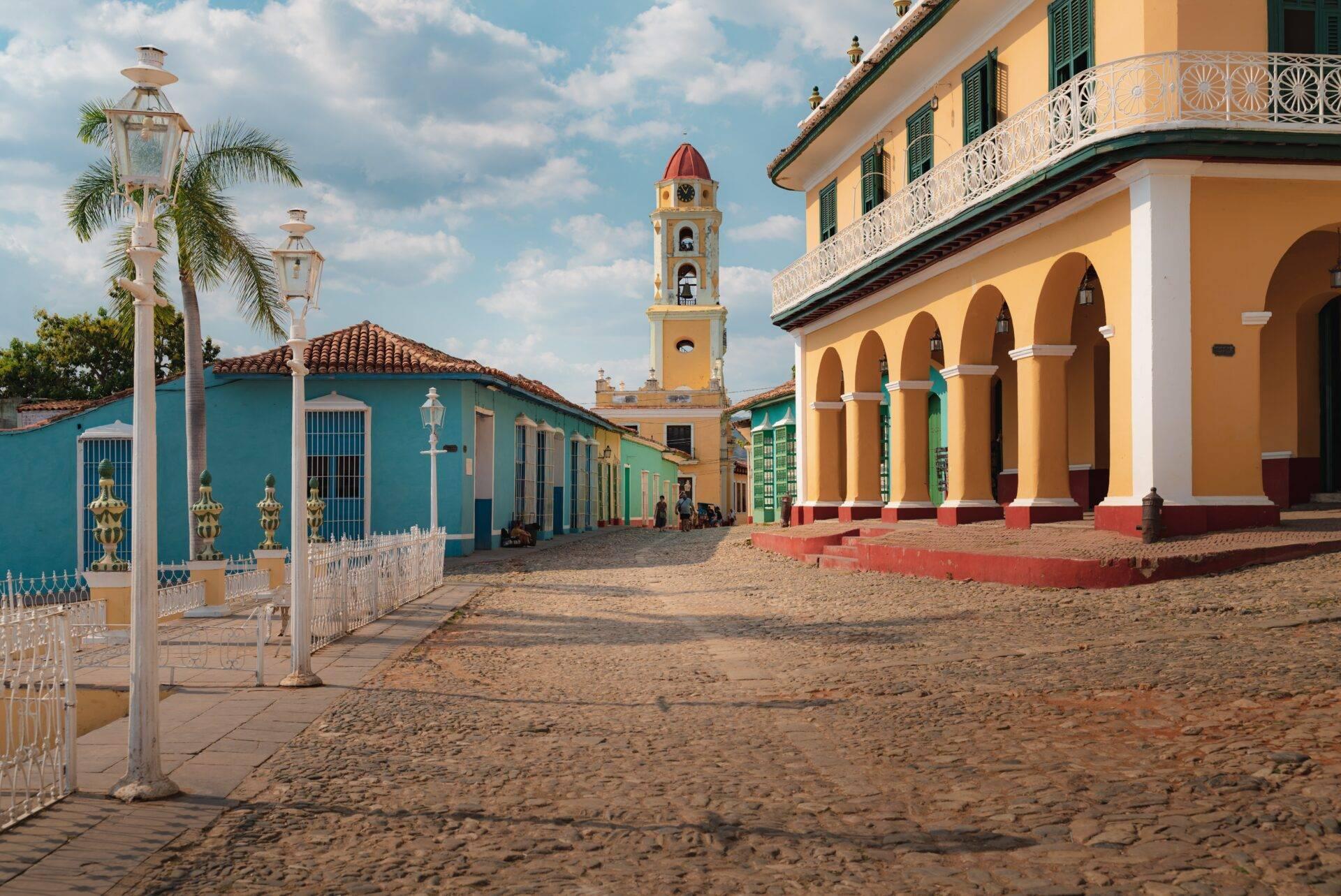 Une place coloniale colorée avec édifices jaunes et bleus, tour avec horloge rouge et arcades, sous ciel dégagé