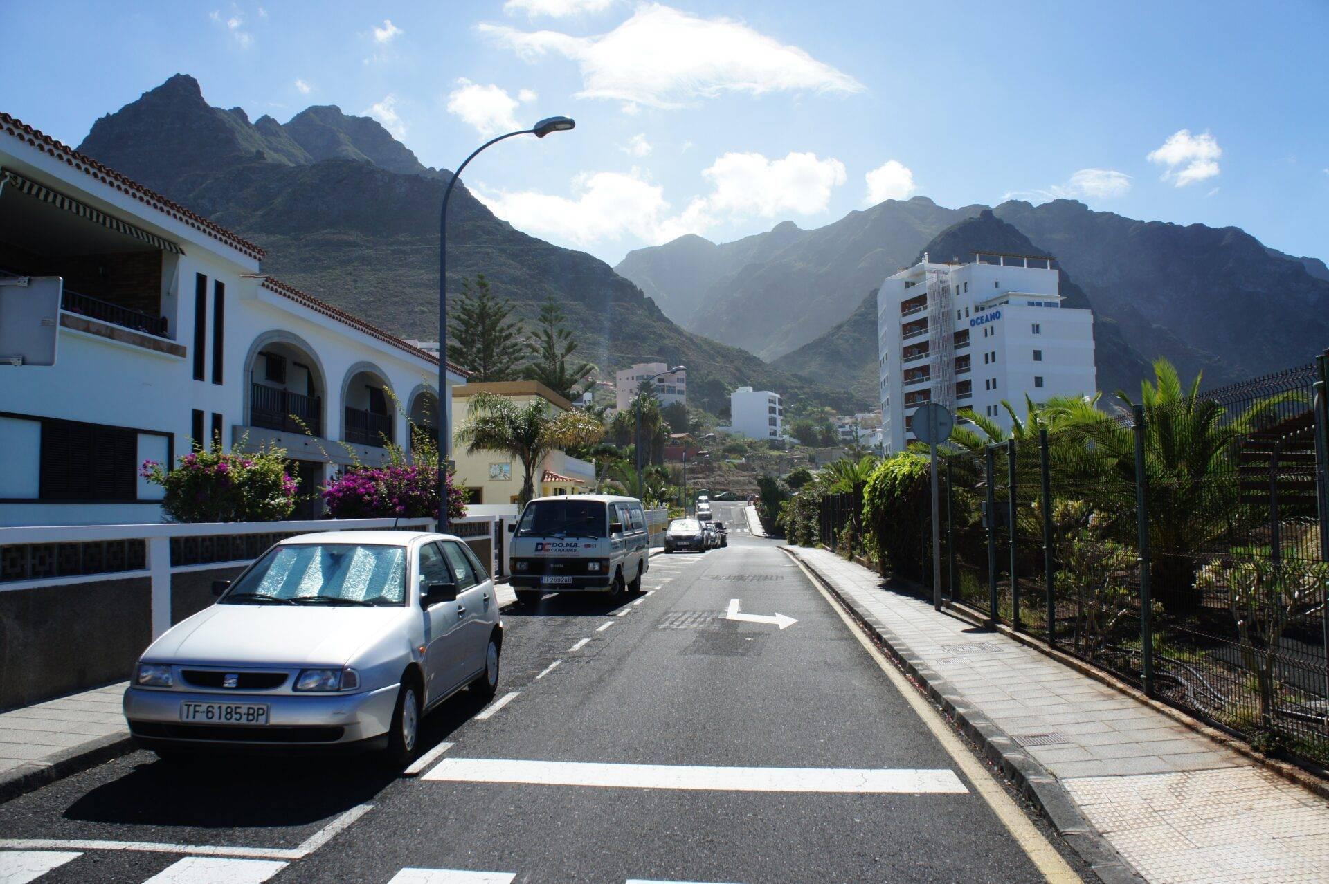 Une rue résidentielle ensoleillée encadrée de montagnes verdoyantes avec immeubles blancs et voitures garées