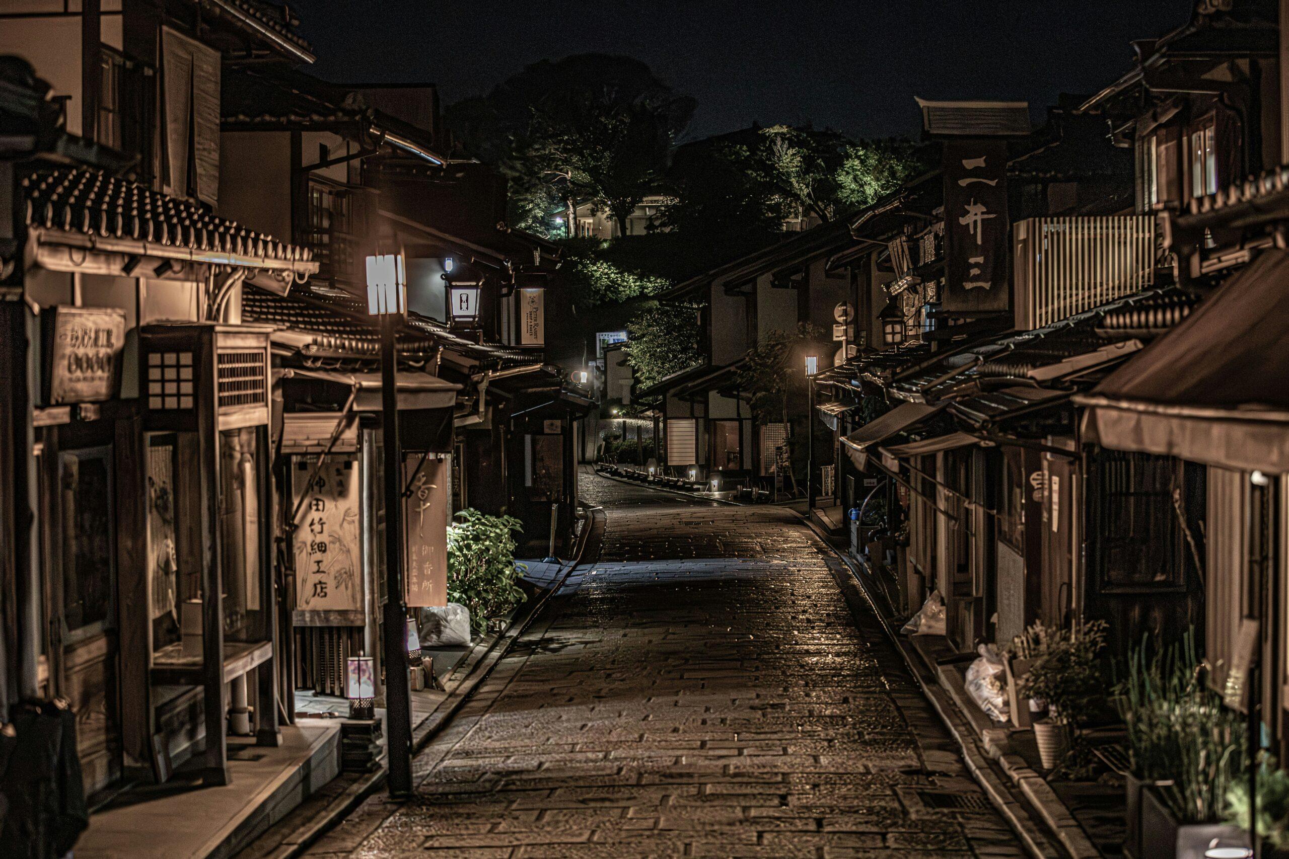 Une ruelle traditionnelle japonaise la nuit, bordée de maisons en bois avec lanternes illuminées et verdure