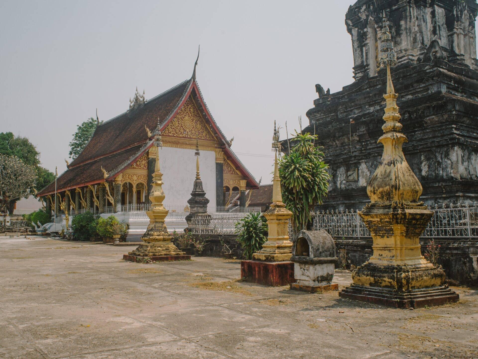 Cour d’un temple bouddhiste laotien avec stupas dorés, bâtiment principal aux toits rouges et ornements dorés