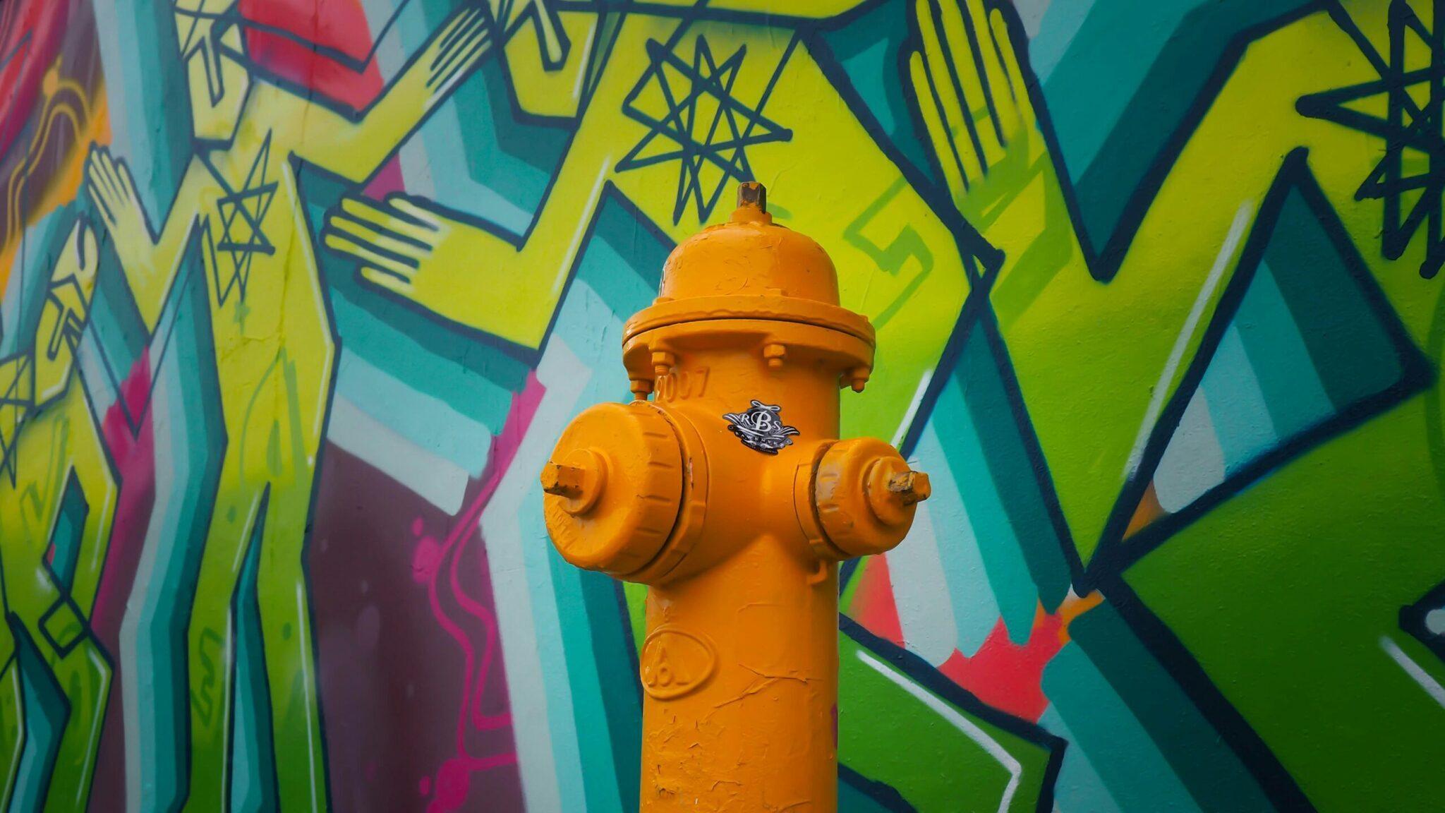 Une borne d’incendie orange vif devant un mur de street art coloré aux motifs géométriques jaunes et turquoise