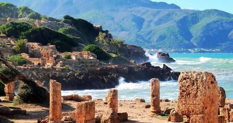 Ruines antiques avec colonnes en pierre face à une plage méditerranéenne aux eaux turquoise et montagnes verdoyantes