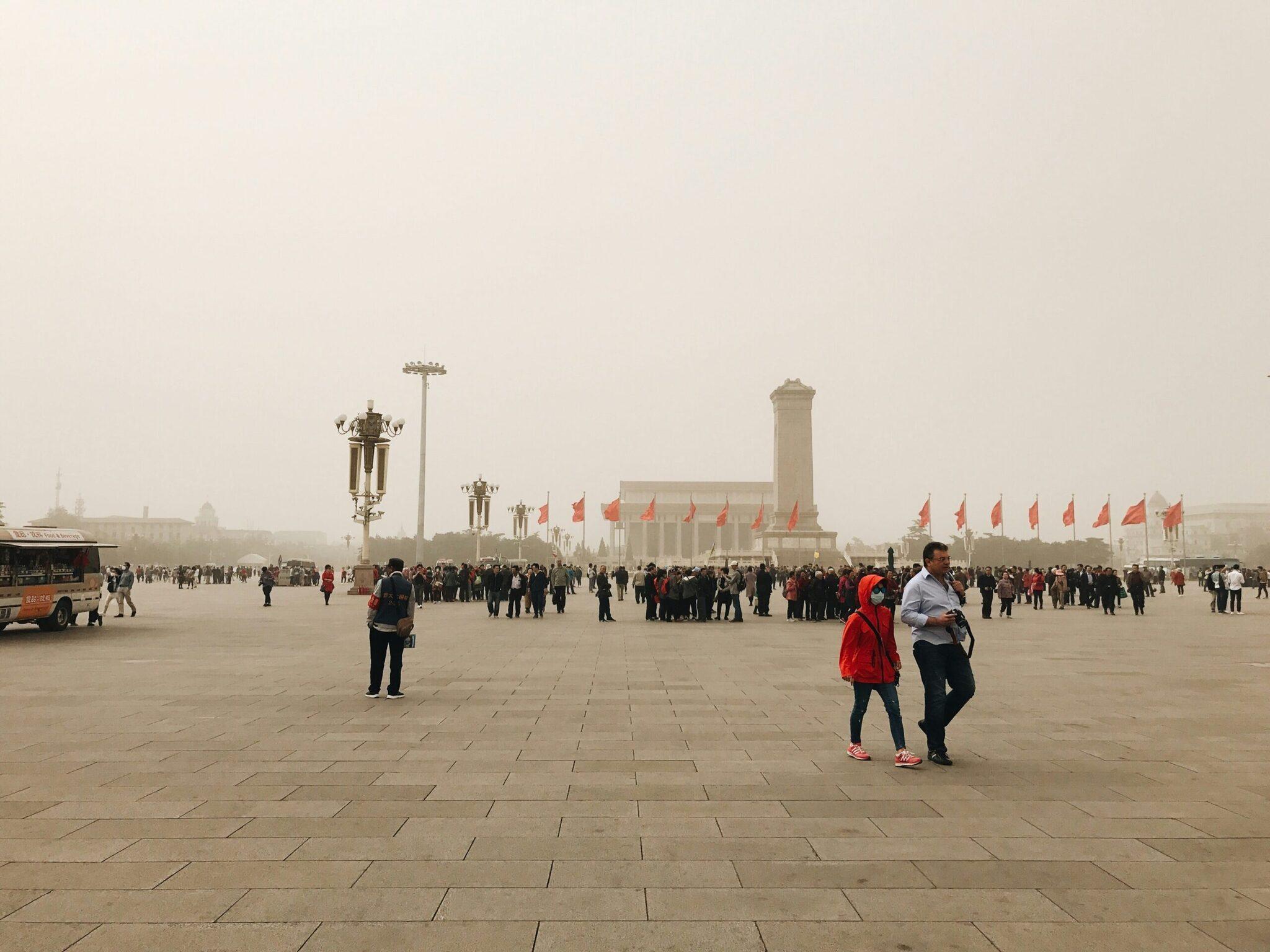 Une vaste place pavée avec des drapeaux rouges, un monument commémoratif et des visiteurs par temps brumeux