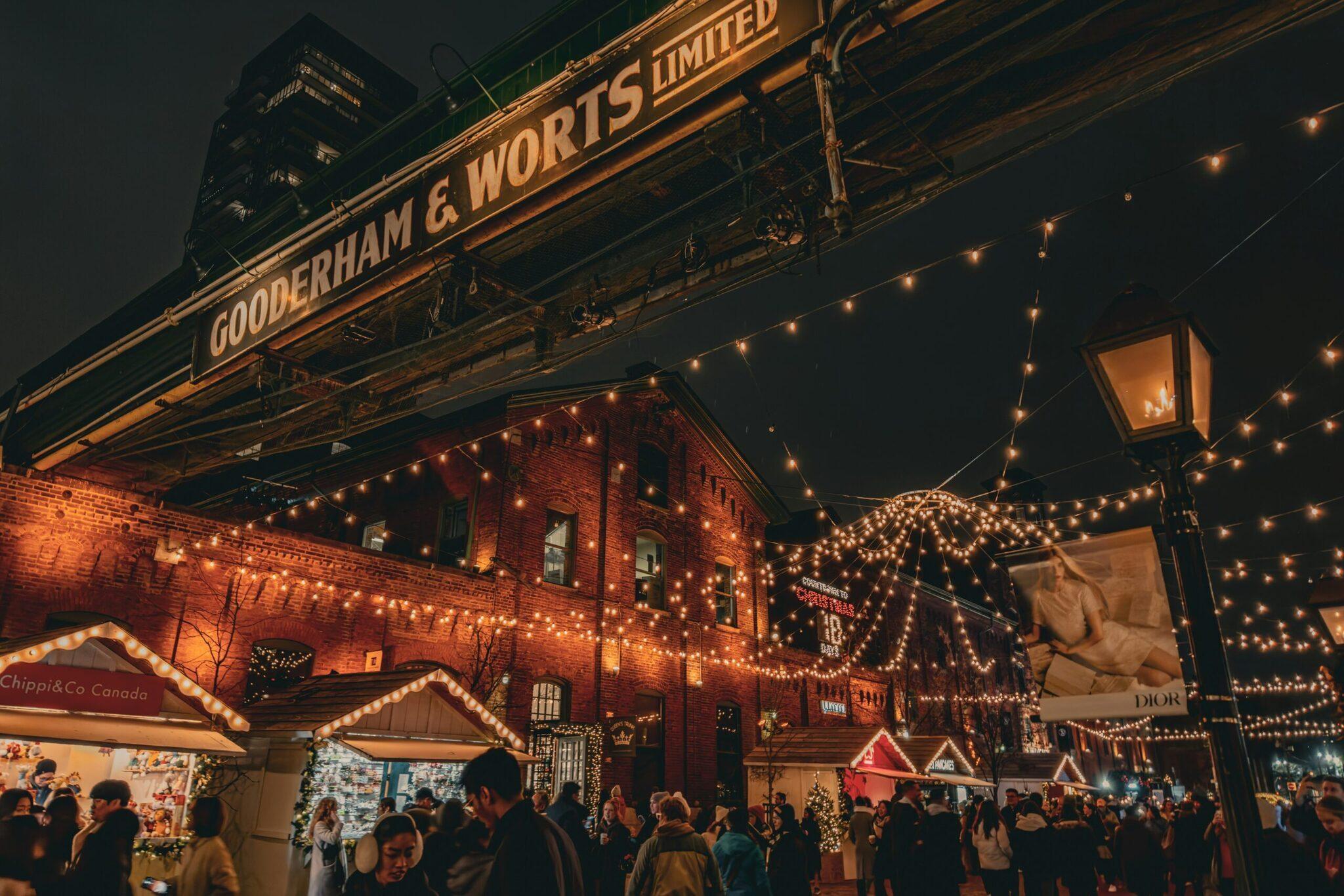 Marché de Noël illuminé avec bâtiments en briques rouges, guirlandes lumineuses et foule de visiteurs en soirée