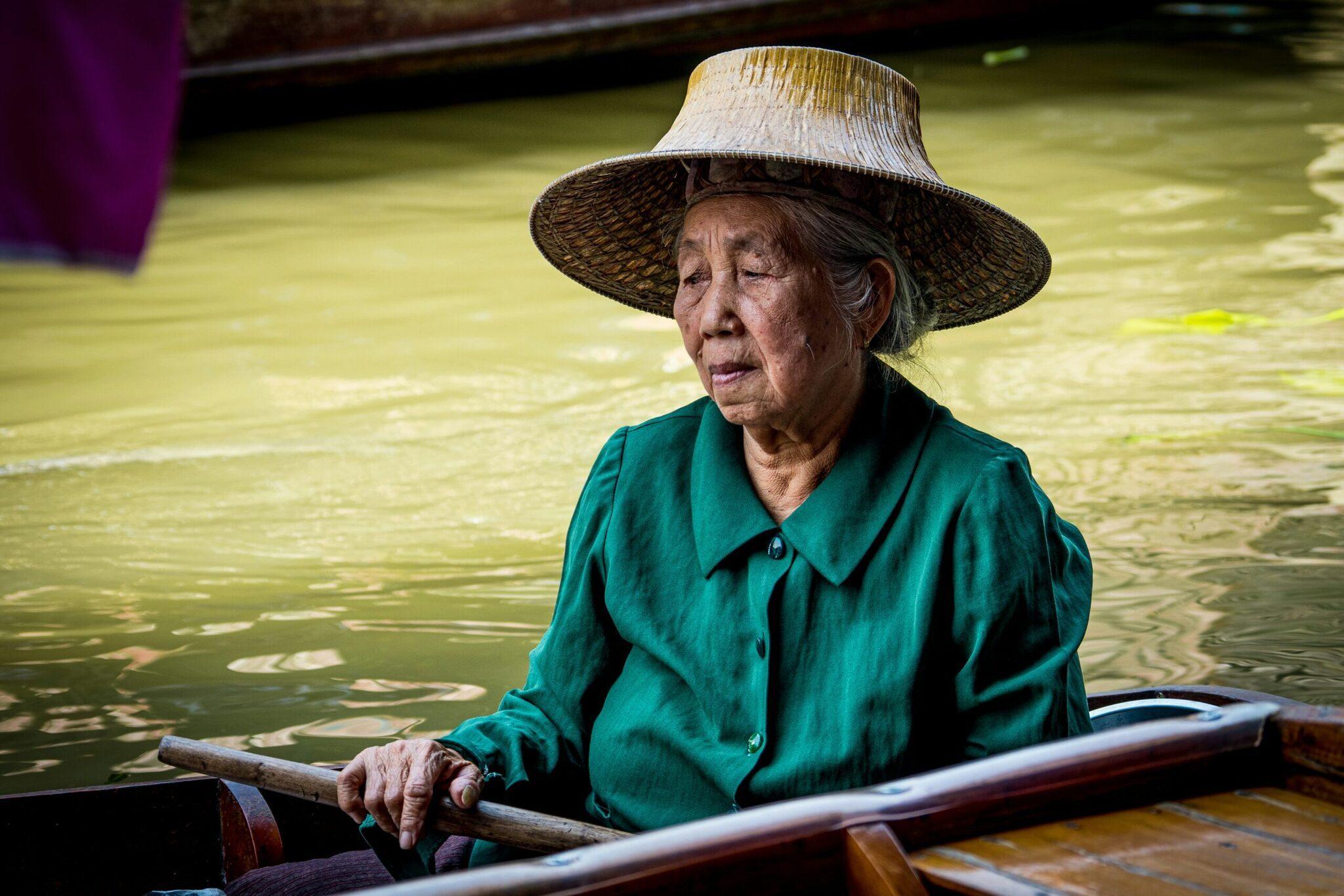 Une femme âgée en chemise verte et chapeau conique traditionnel navigue en bateau sur un canal aux eaux vertes