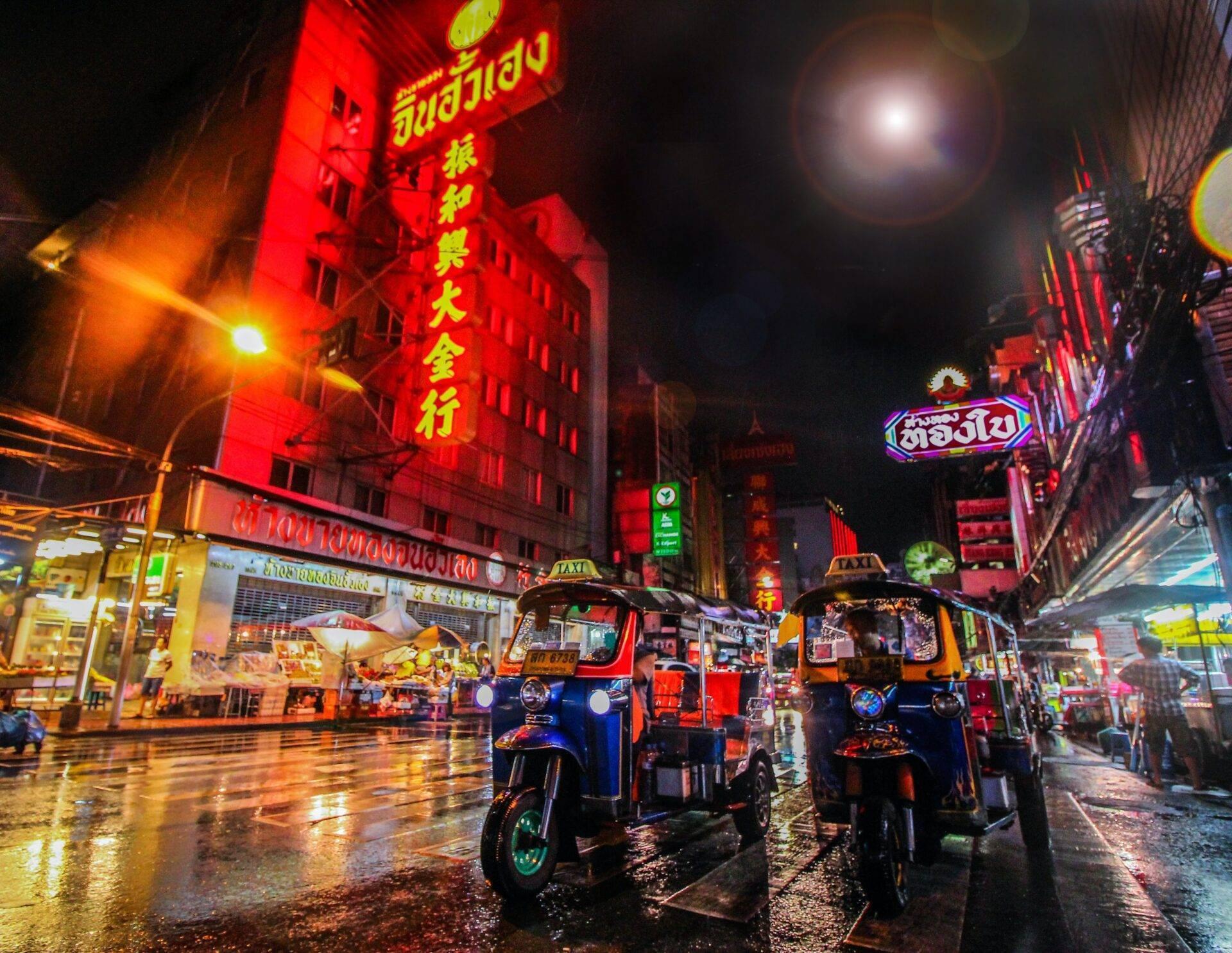 Rue animée de nuit en Thaïlande avec tuk-tuks bleus, enseignes lumineuses rouges et jaunes, sol mouillé reflétant les lumières