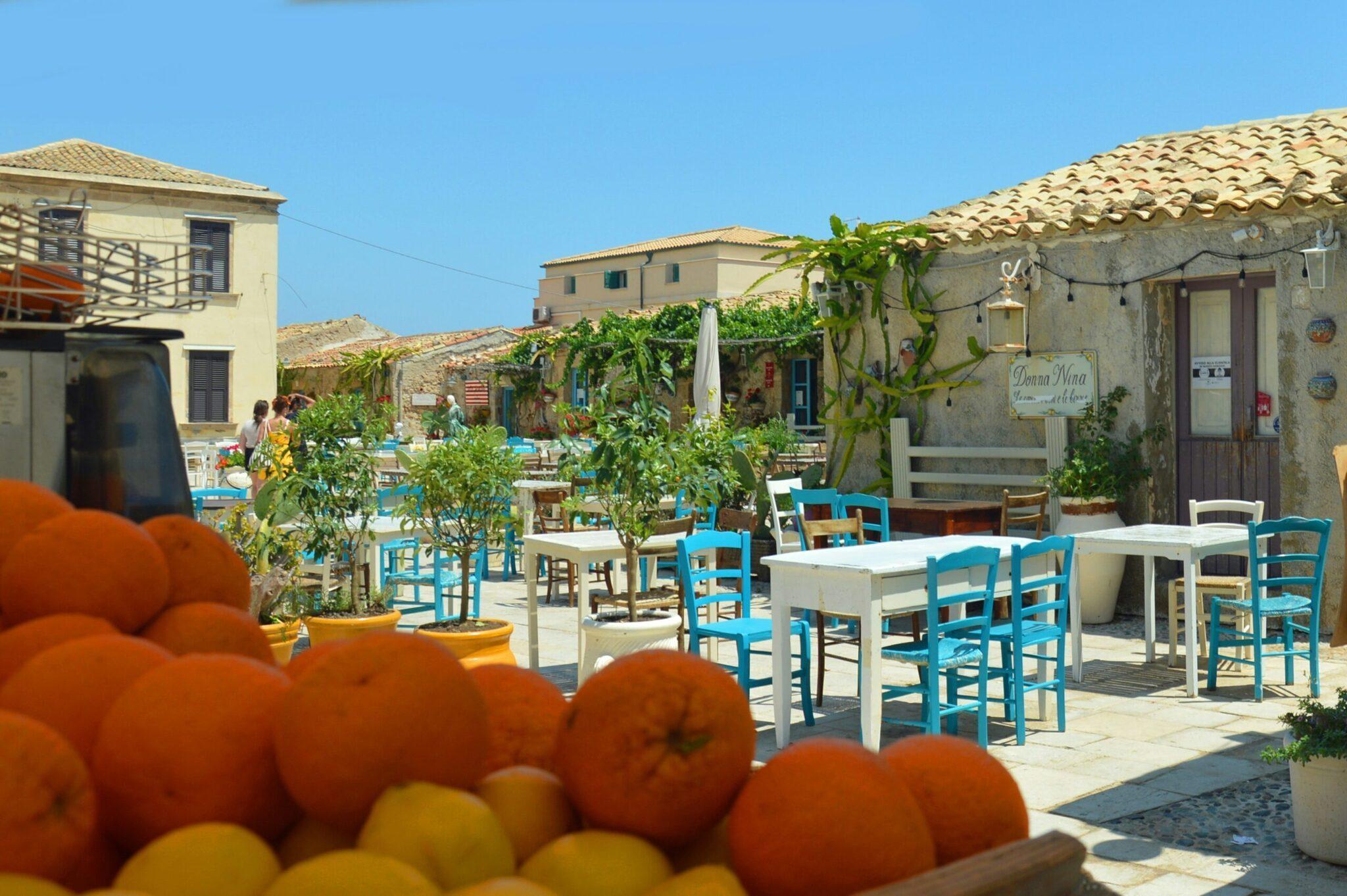 Une terrasse méditerranéenne avec tables bleues et blanches, agrumes frais au premier plan, village sicilien typique en arrière-plan