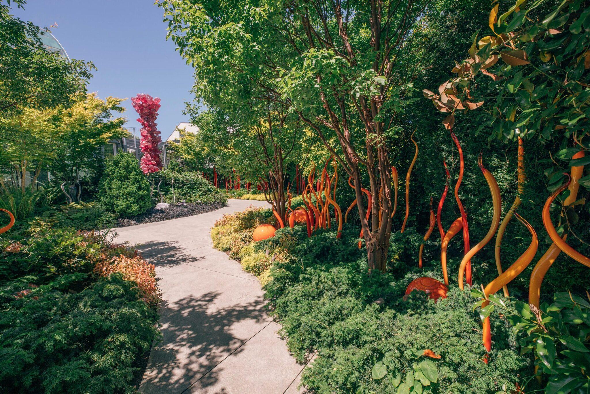 Allée de jardin moderne bordée d’arbres aux troncs peints en rouge et orange vif, avec sculptures colorées