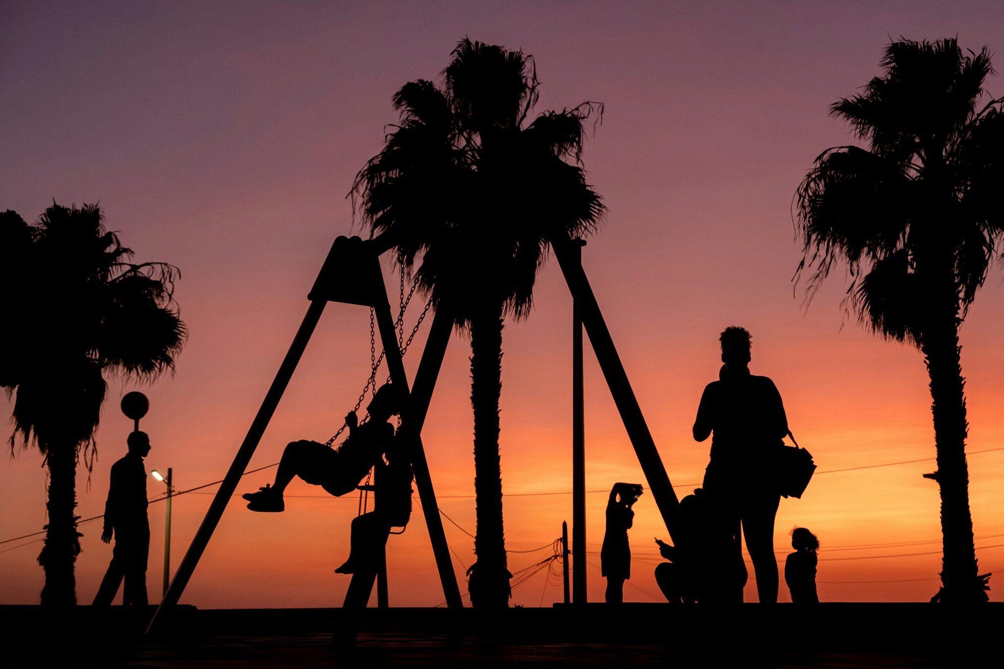 Silhouettes d’enfants jouant sur une balançoire au coucher de soleil avec palmiers et ciel orange