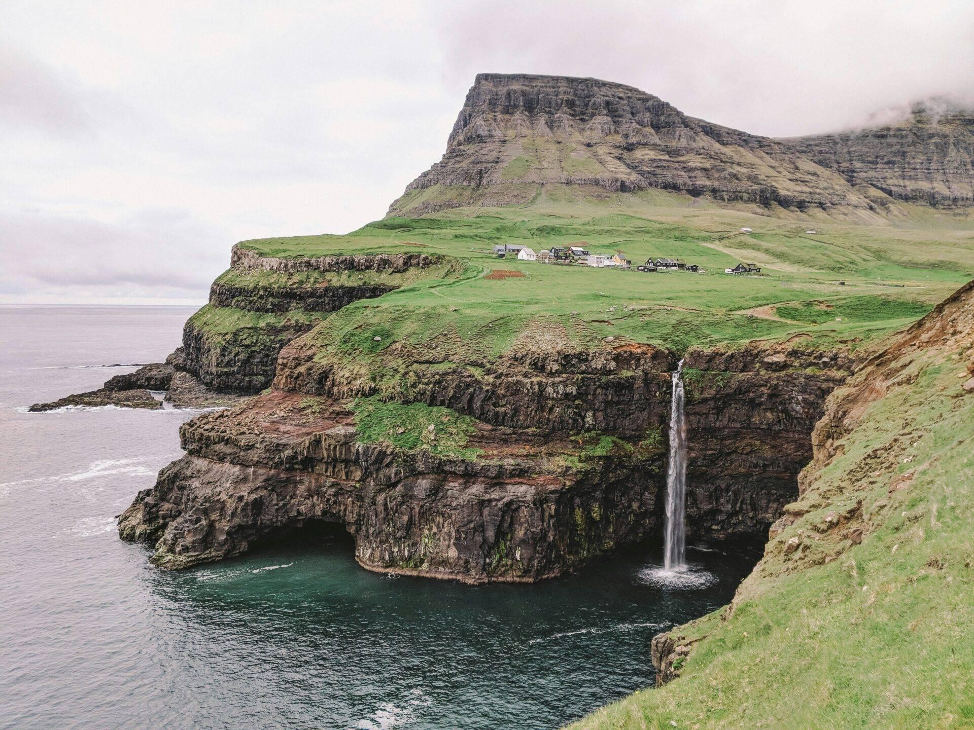 Paysage côtier montagneux avec cascade, falaises verdoyantes et fermes isolées sous ciel nuageux