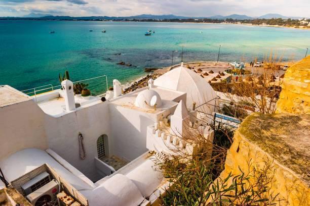 Maison blanche méditerranéenne perchée sur une falaise rocheuse surplombant une mer turquoise avec bateaux