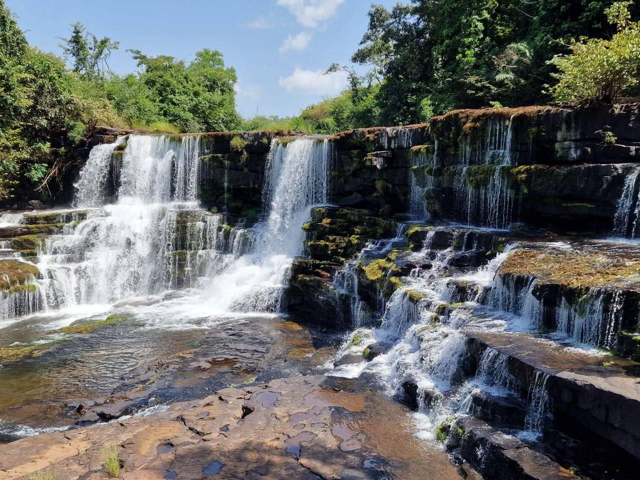 Une cascade spectaculaire multi-étages avec roches mousseuses, entourée de végétation luxuriante et d’un ciel dégagé