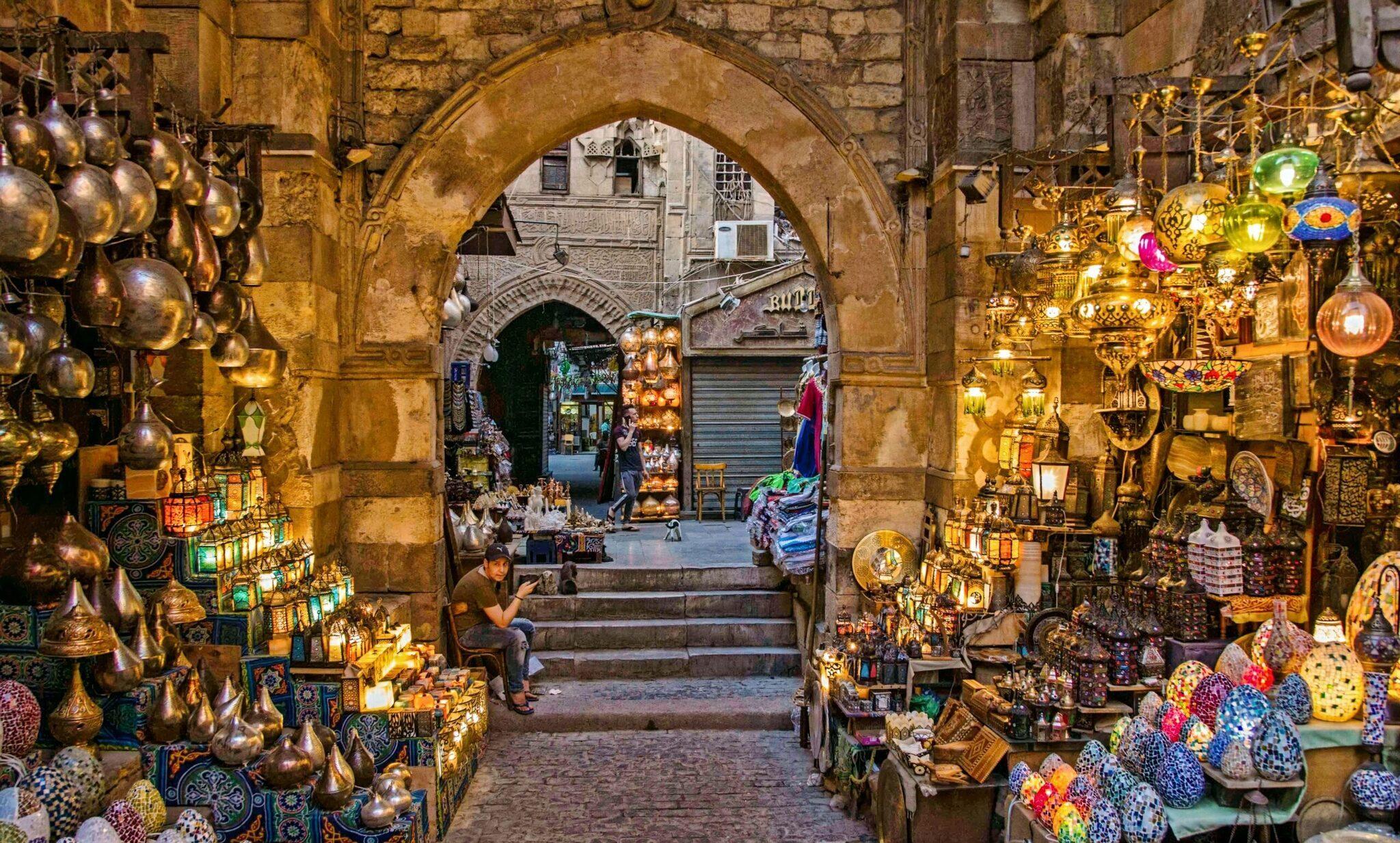 Une ruelle couverte d’un marché oriental illuminée, bordée de lanternes dorées et d’objets artisanaux colorés, avec une arche de pierre