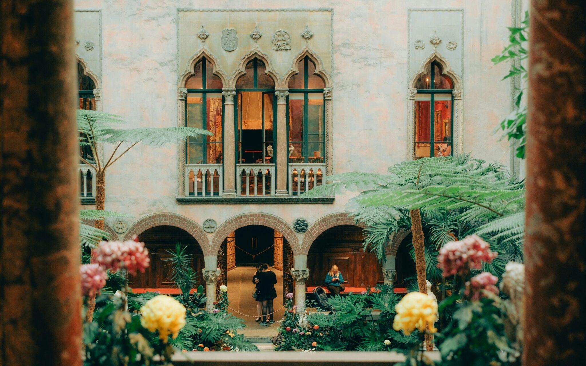 Cour intérieure avec arcades en brique, fenêtres gothiques, plantes tropicales et personnes flânant dans une atmosphère méditerranéenne