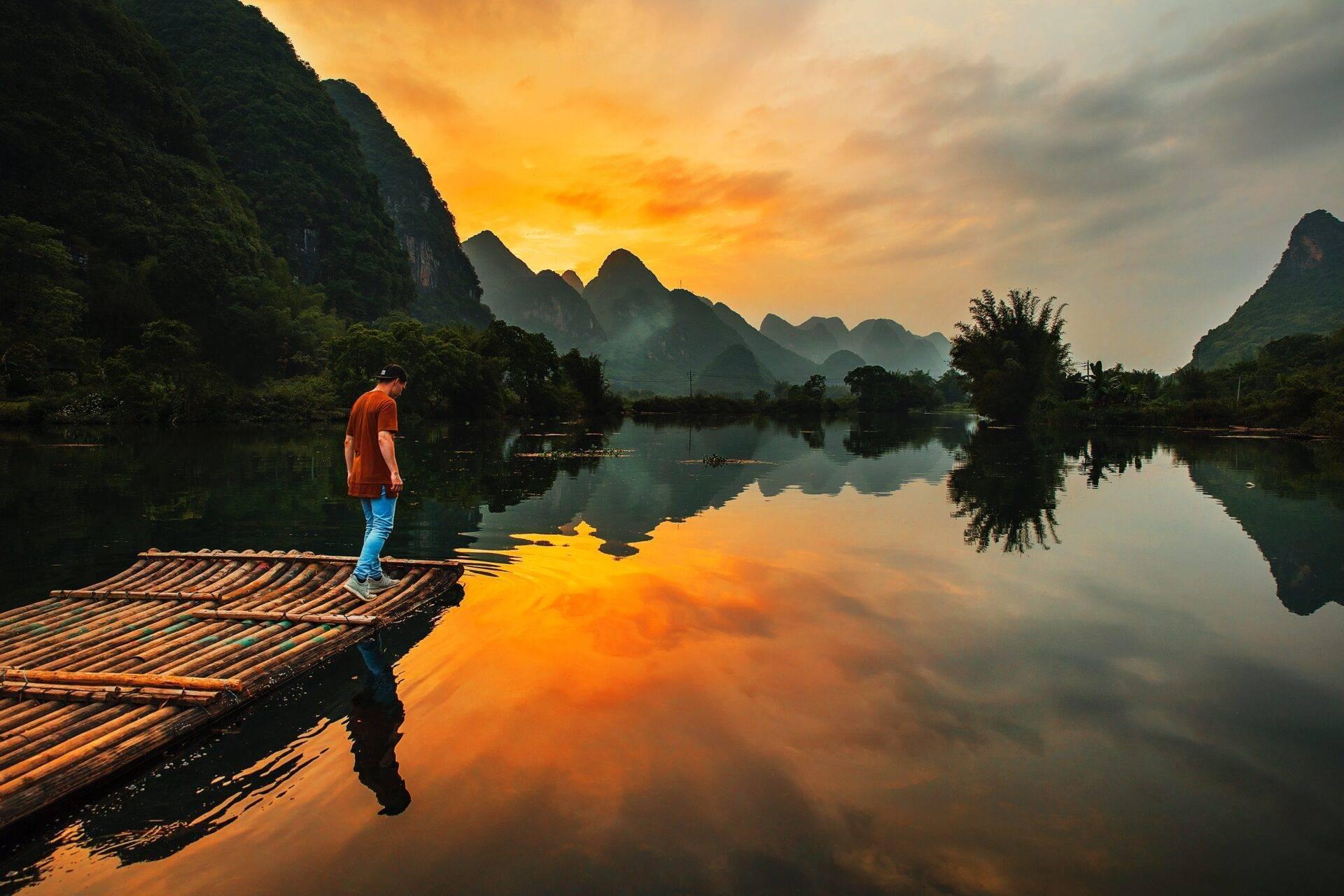 Un homme en chemise orange sur un radeau de bambou contemple une rivière calme entourée de montagnes karstiques au coucher du soleil