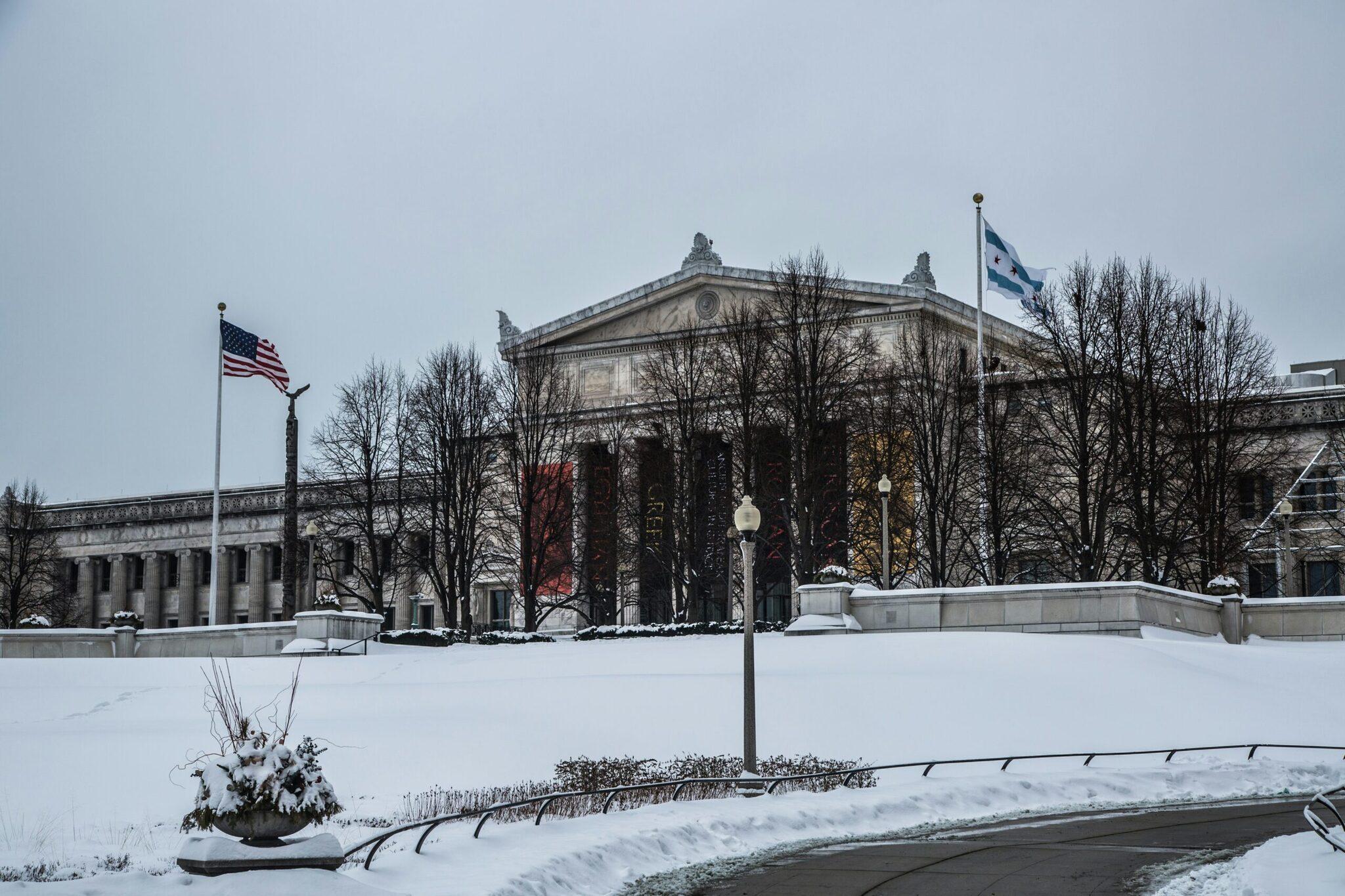 Un bâtiment néoclassique enneigé avec colonnes et drapeau américain, entouré d’arbres nus en hiver