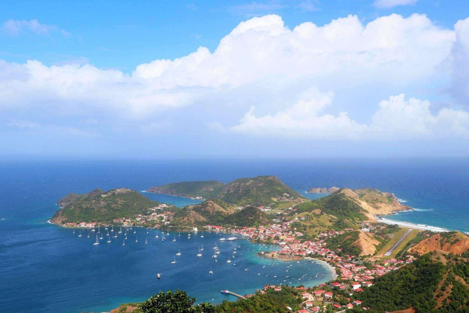 Vue aérienne d’une baie tropicale avec voiliers ancrés, plages de sable blanc et végétation luxuriante sur les collines côtières