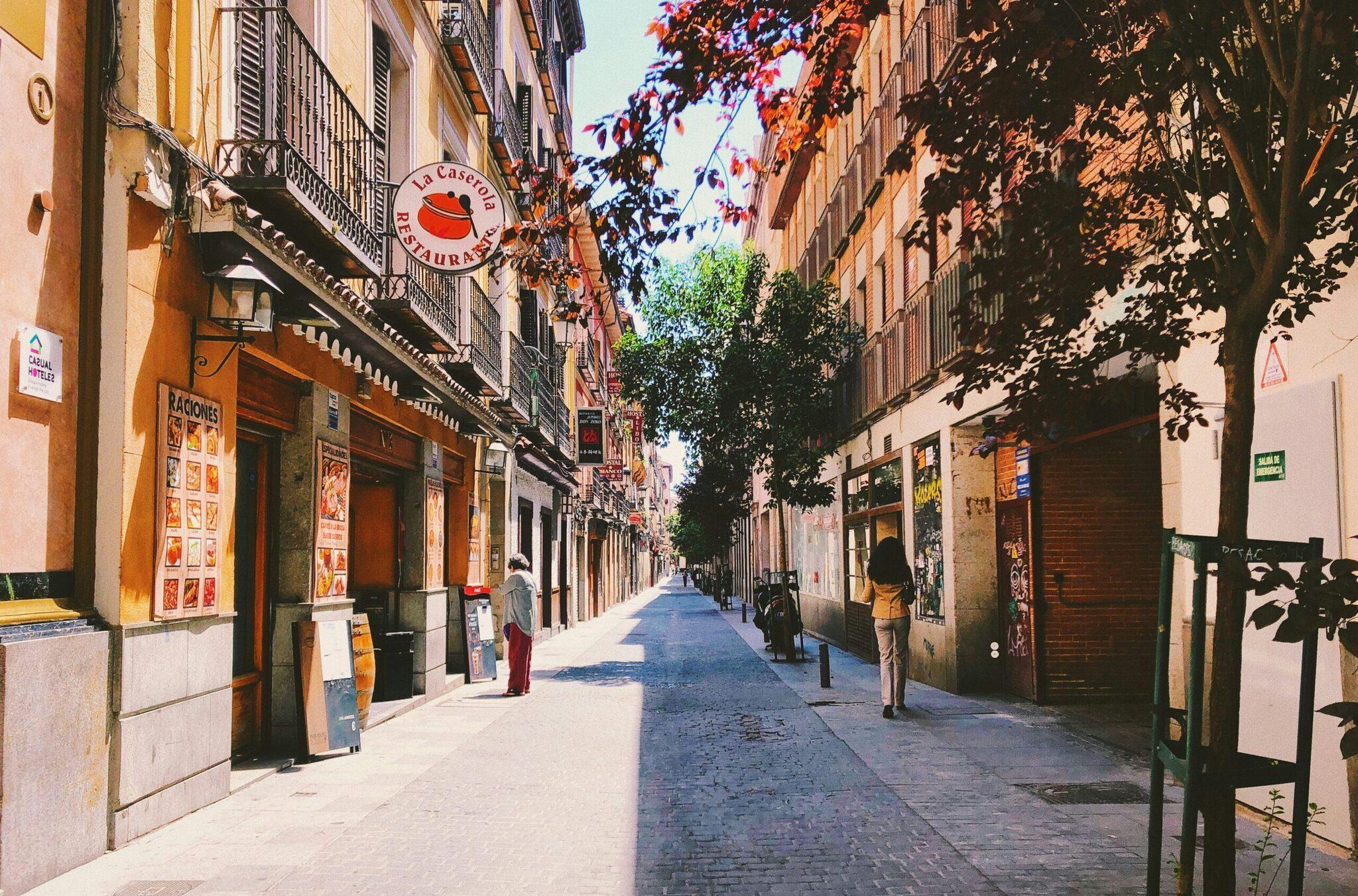 Une rue étroite pavée d’une ville espagnole avec façades ocre, commerces locaux et verdure, sous un ciel ensoleillé