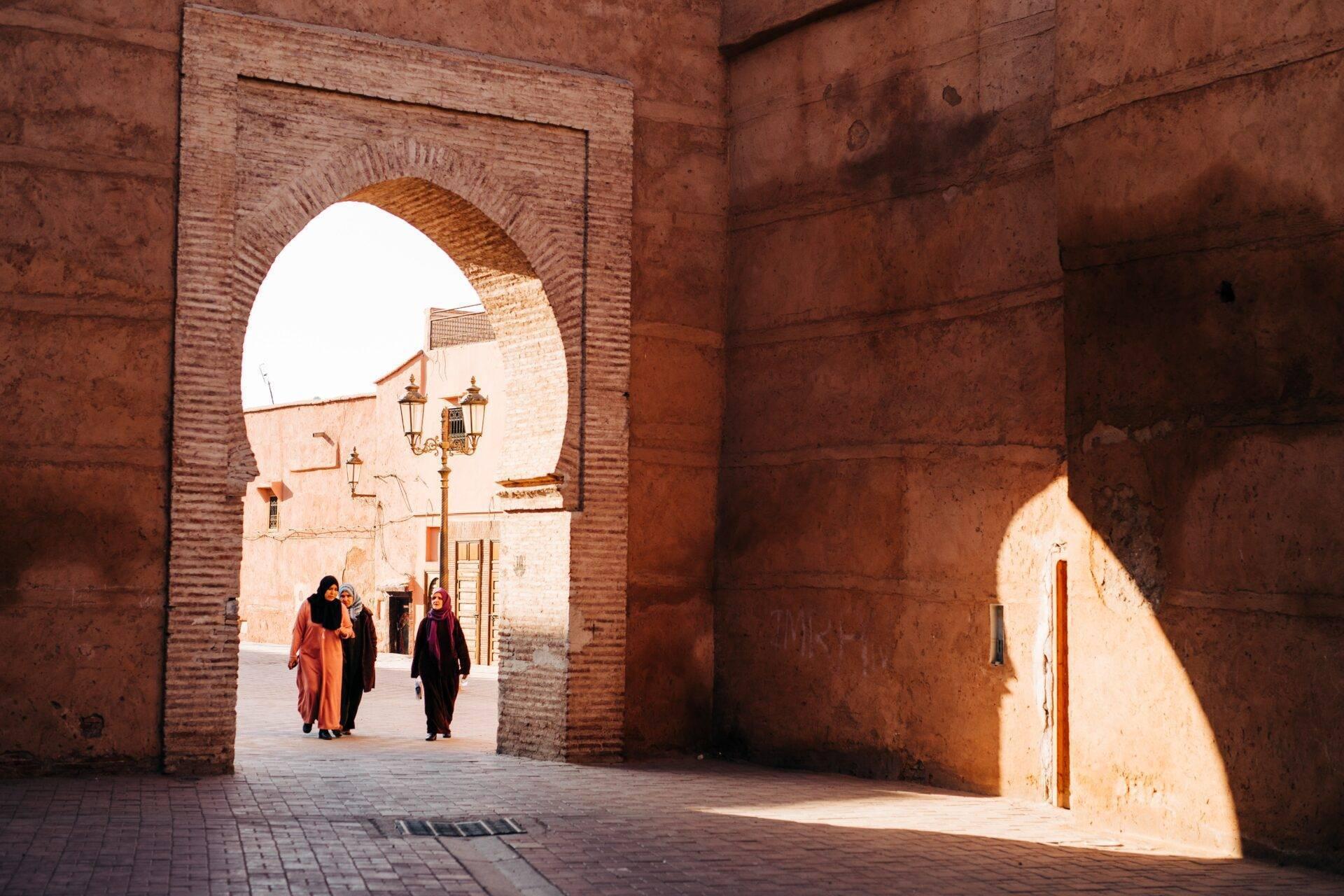 Trois femmes traversent une arche en brique rouge dans une medina marocaine baignée de lumière naturelle