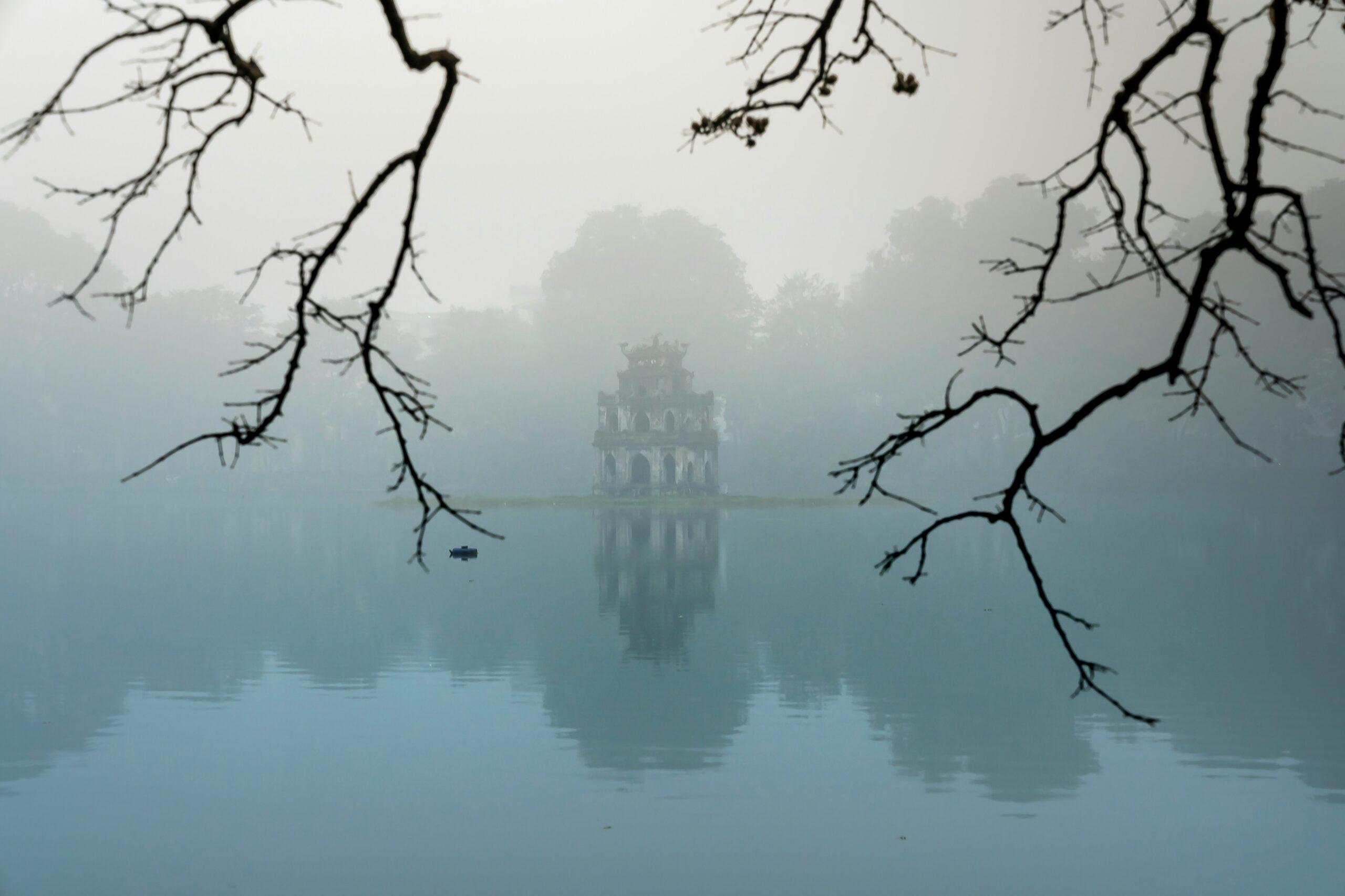 Une tour historique se reflète dans un lac calme sous un brouillard épais, encadrée par des branches dénudées