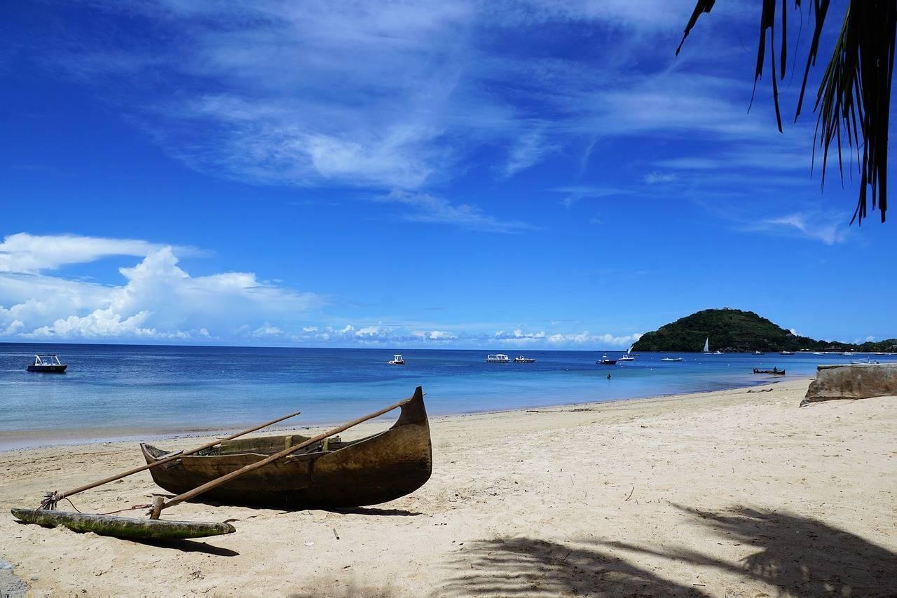 Une barque traditionnelle échouée sur une plage de sable blanc face à une baie turquoise et une île boisée